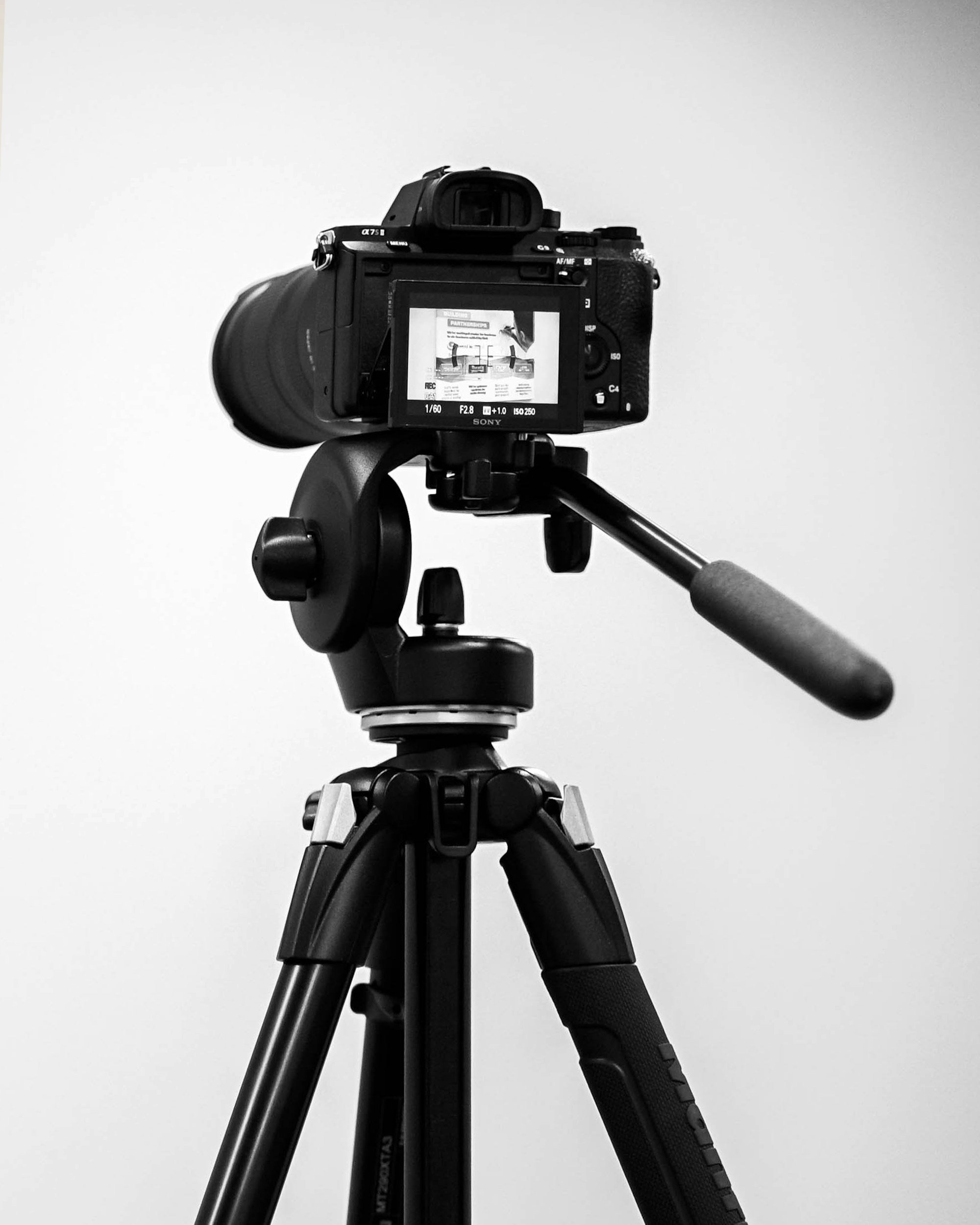 A camera mounted on a tripod, with the camera screen displaying a preview of a presentation or image, all in black and white.