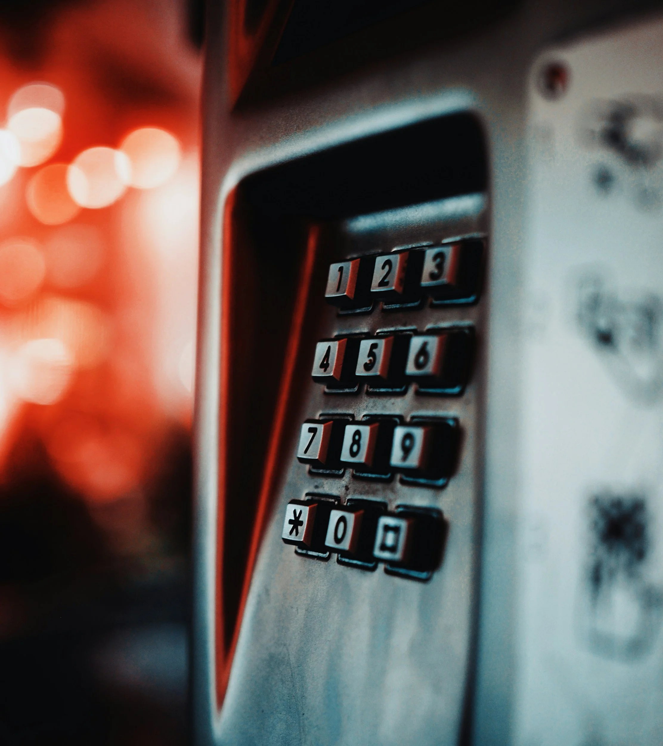 Close-up of a telephone keypad with illuminated number keys 1 to 9, 0, and symbols star and pound, on an outdoor payphone, with soft orange bokeh lights in the background.