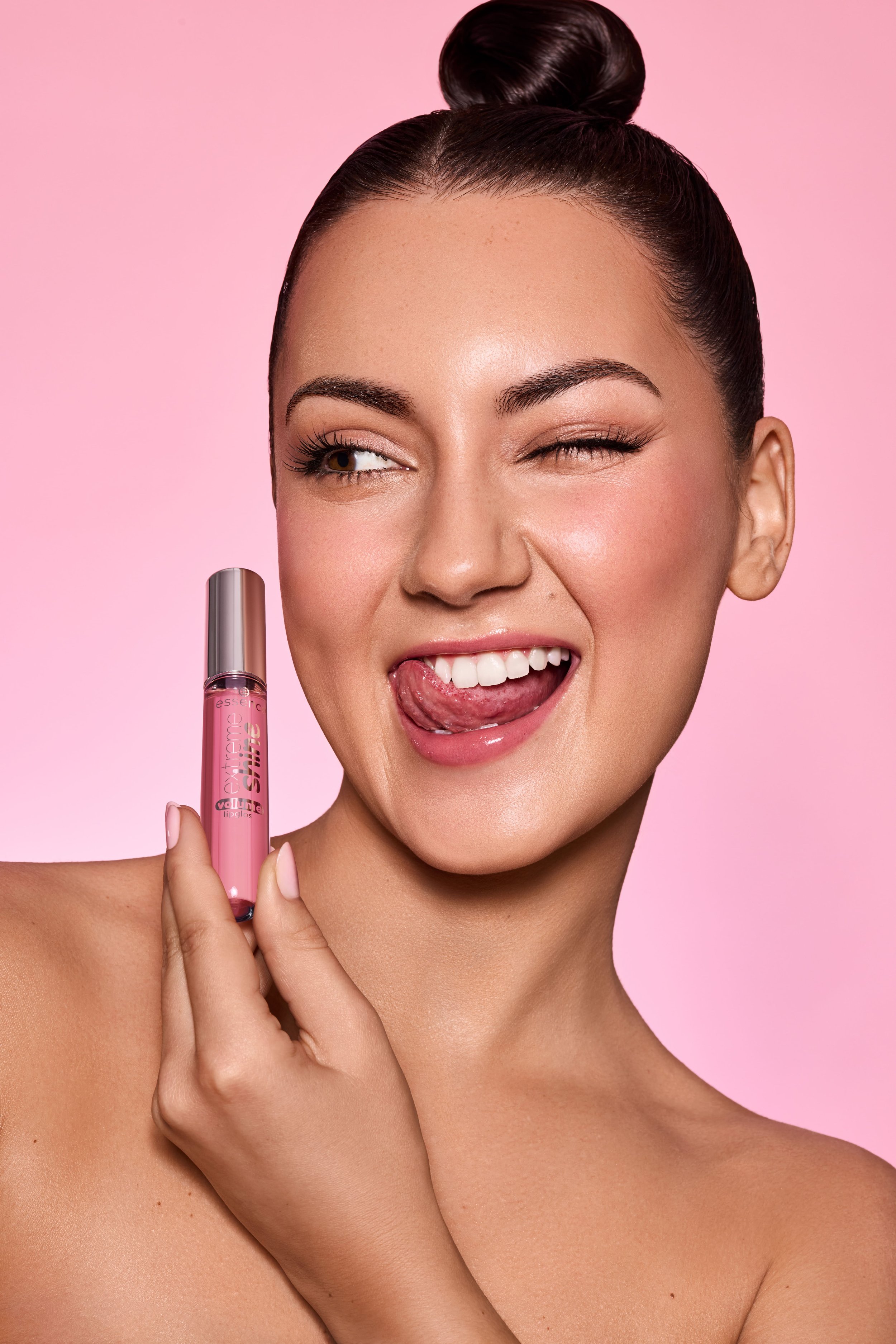 Eine lachende Frau mit dunklem Haar in einem hohen Dutt zeigt einen pinken Lippenstift in der Hand. Sie hat einen pinken Hintergrund.