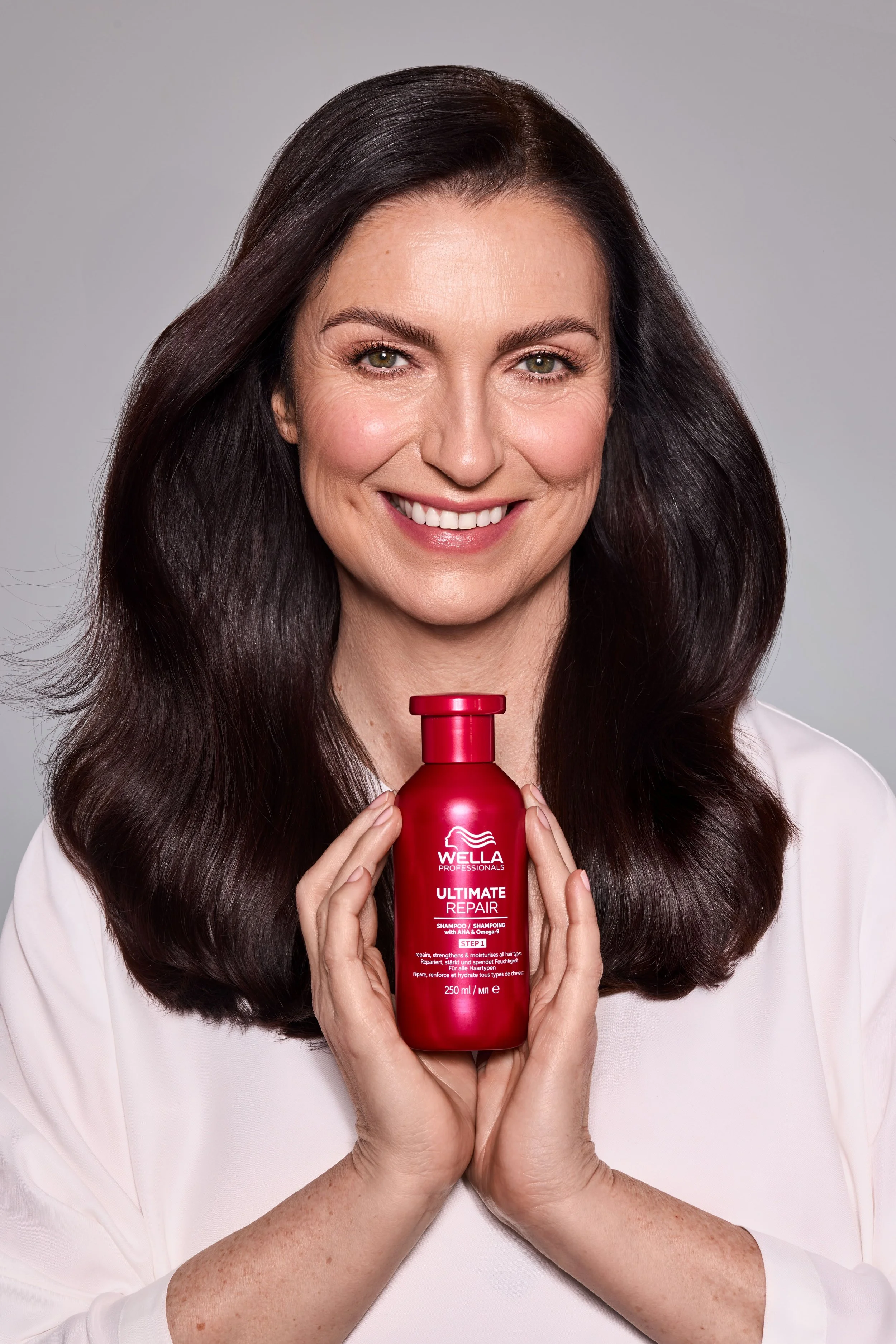 Eine lachende Frau mit braunen, langen Haaren hält eine rote Flasche Wella Ultimate Repair Shampoo vor ihrer Brust. Sie trägt eine weiße Bluse vor einem grauen Hintergrund.