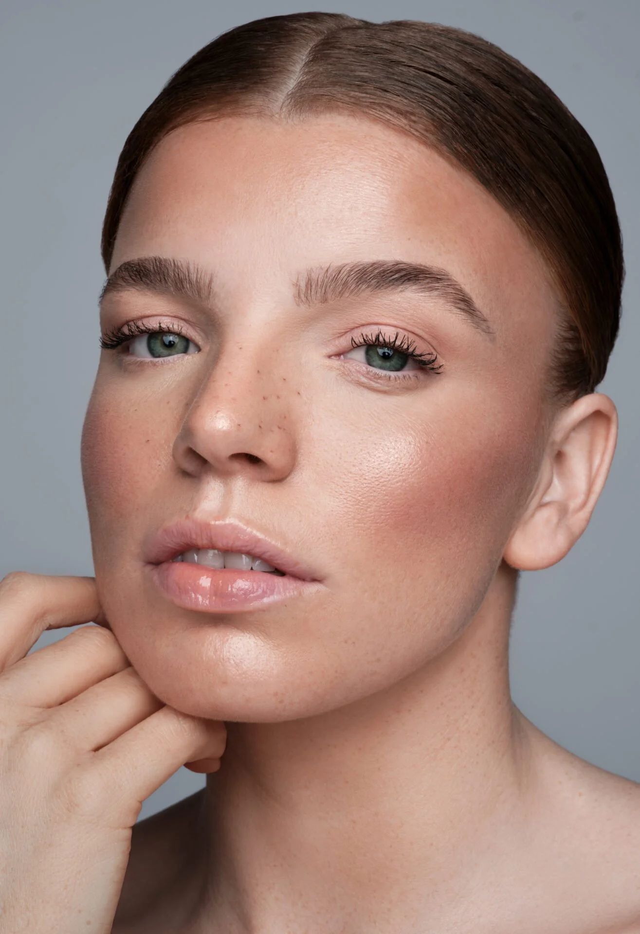 Nahaufnahme eines jungen Mädchens mit glatter Haut, braunen Haaren, blauen Augen und natürlichen makeup, das sanft an seinem Kinn hält.