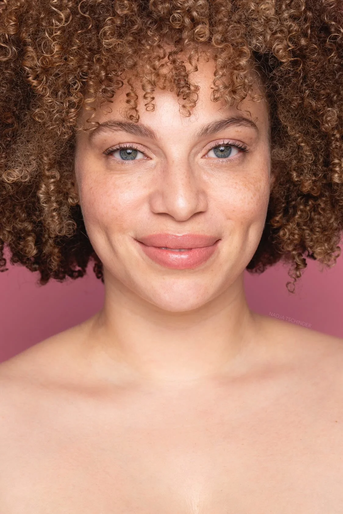 Nahaufnahme eines lachenden Mädchens mit lockigem braunem Haar vor rosa Hintergrund.