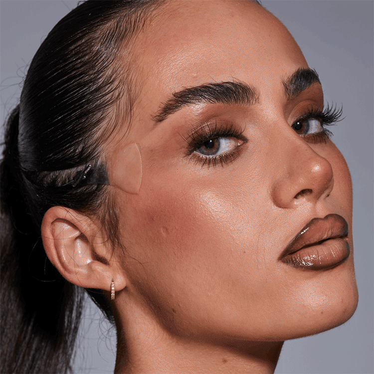 Nahaufnahme eines Mädchens mit dunklem, glattem Haar, geschminkten Augen, Lippen und Augenbrauen, in einem Studio mit grauem Hintergrund.