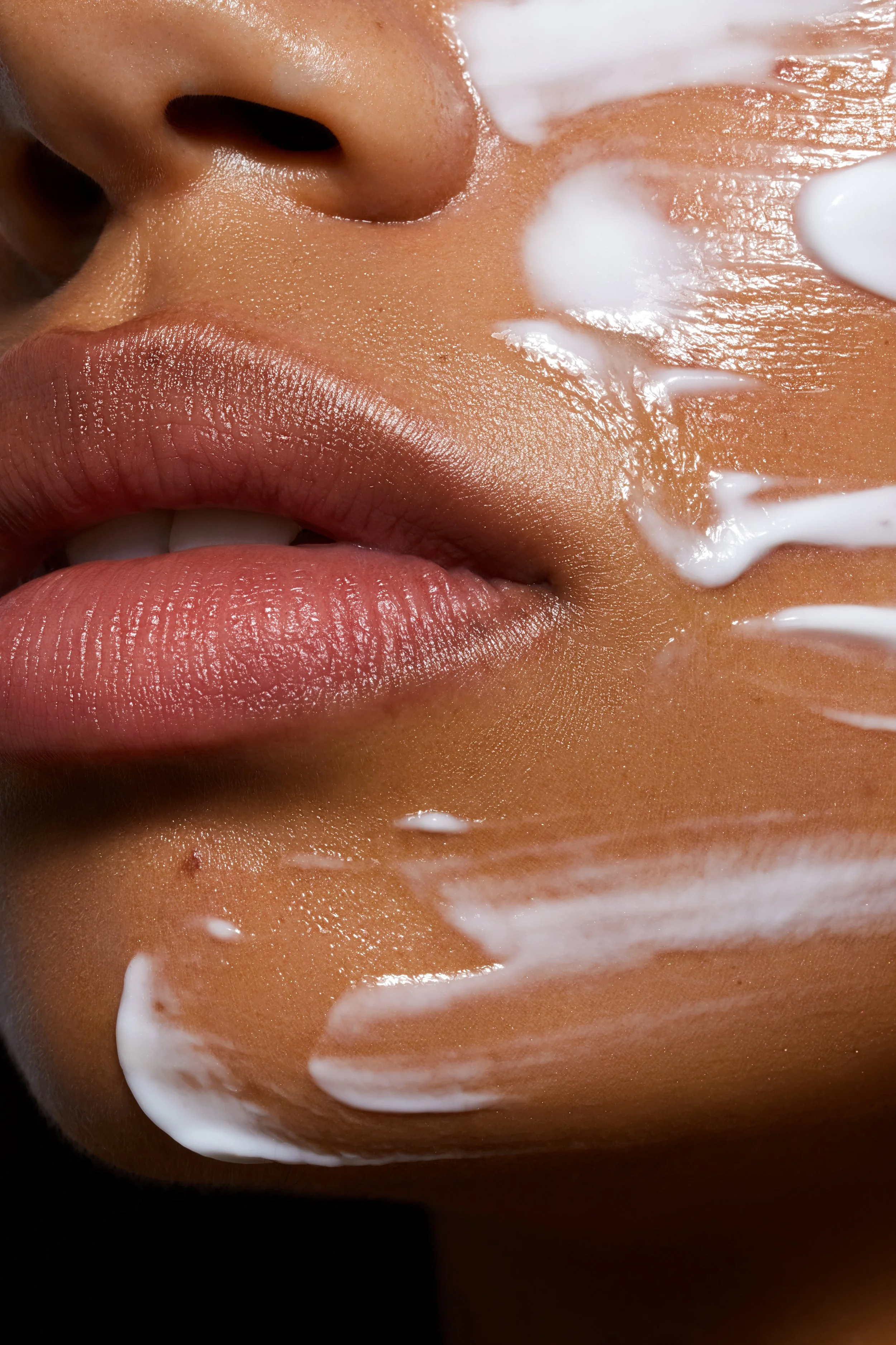 Nahaufnahme eines Gesichts mit fokus auf Lippen, Nase und Wange, während auf der Haut eine weiße Creme verteilt wird.