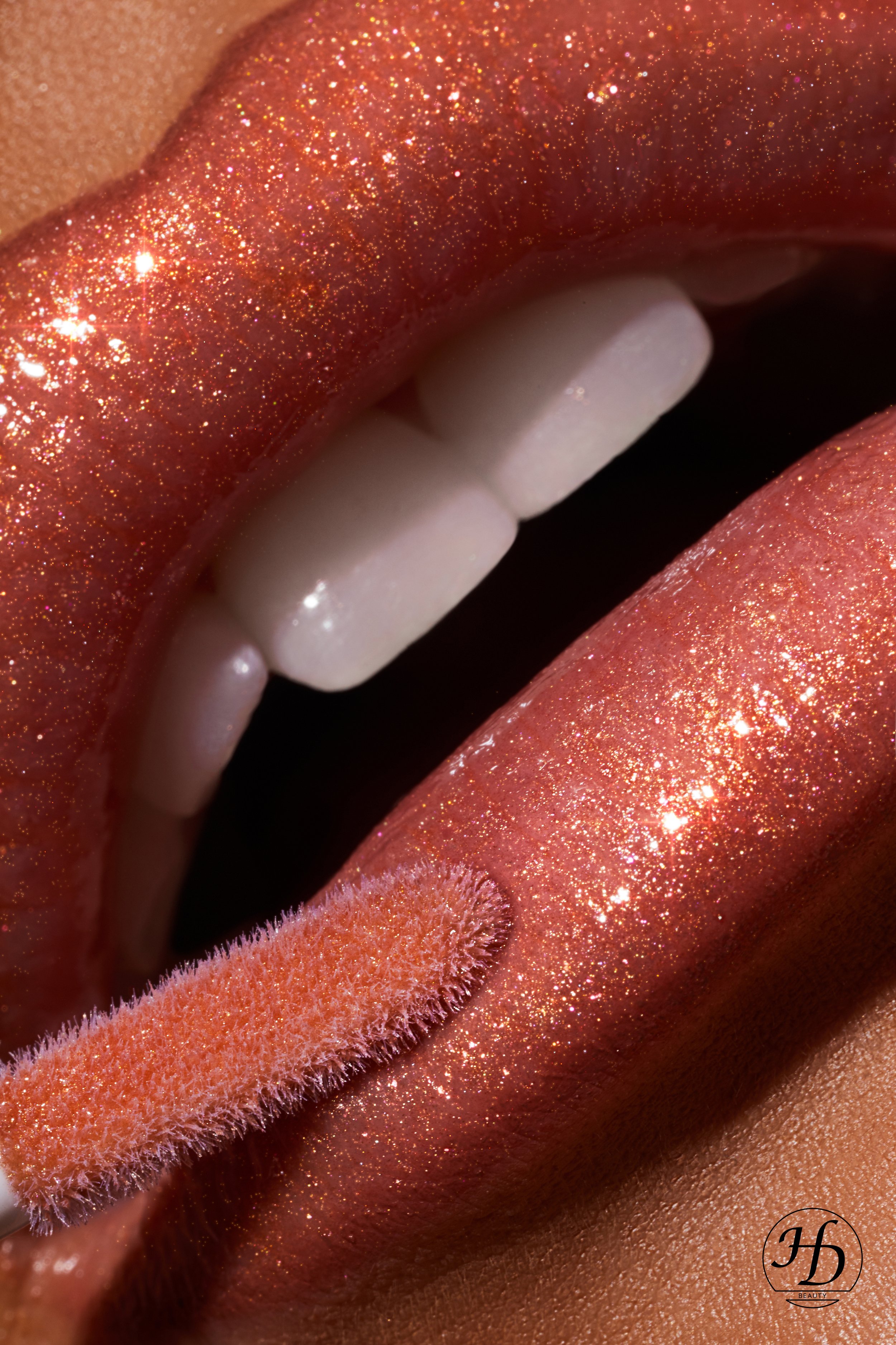 Nahaufnahme von glitzernden, rosegoldfarbenen Lippen mit offenem Mund, sichtbar mit weißen Zähnen und einer Lippenstift-Applikator. Das Bild betont die glitzernde Textur der Lippen. © HD Beauty