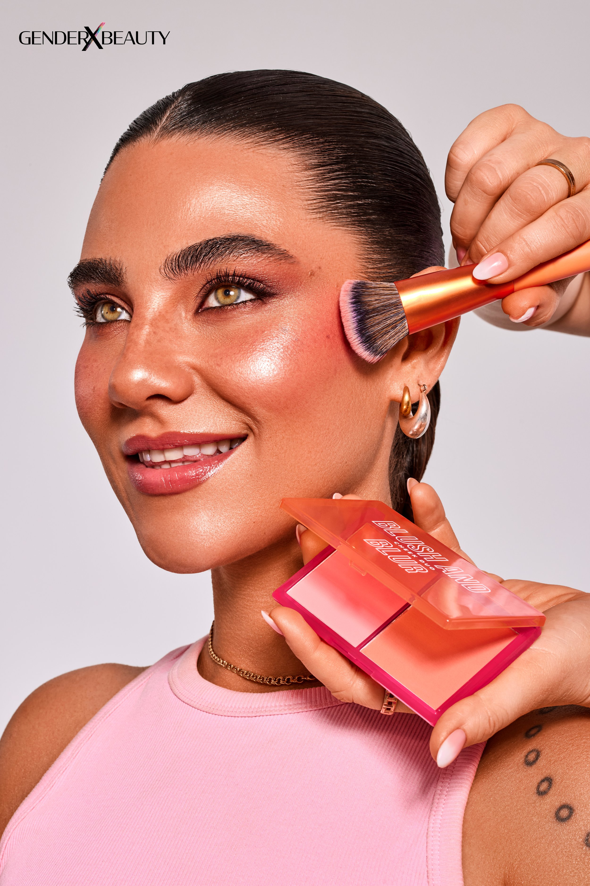 Eine Frau mit dunklem, glatt zurückgekämmtem Haar wird geschminkt. Eine Hand hält eine Puderpinsel und tupft auf ihre Wange, während eine andere Hand eine rosafarbene Palette mit dem Text "Push Said Blush" hält.