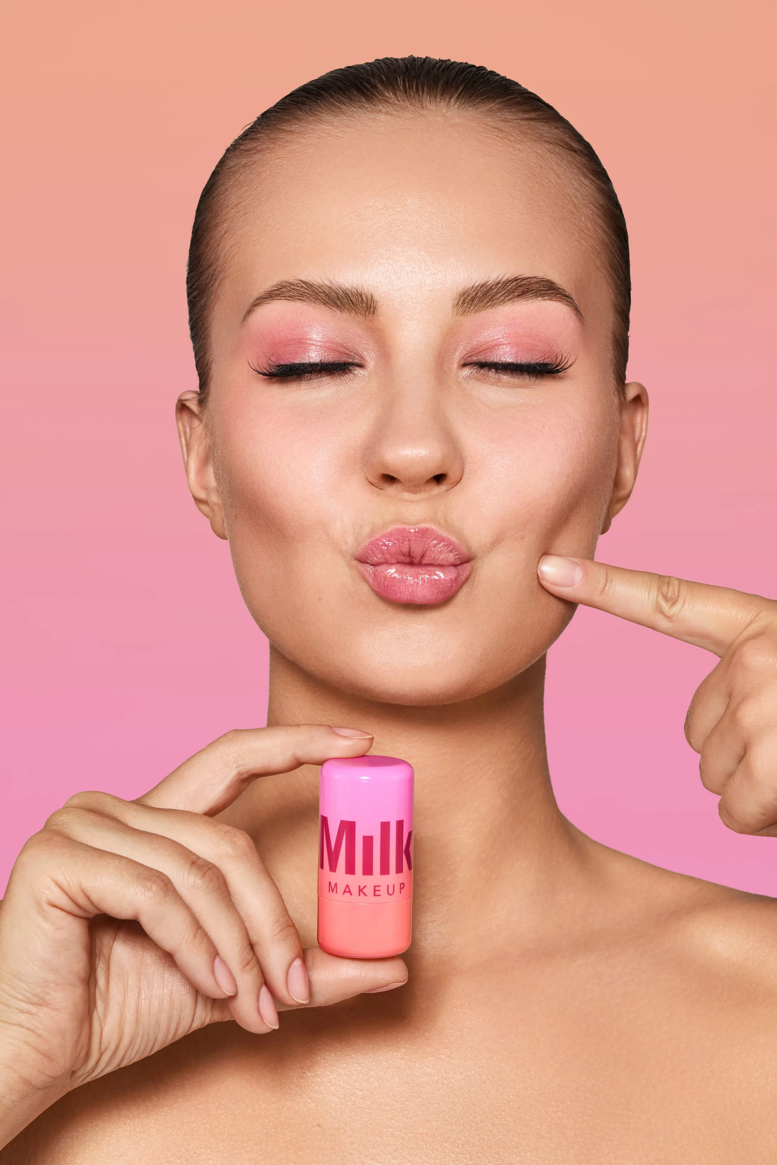 Junge Frau mit geschlossenen Augen und Lippen, die eine pinke Lippenpflege hält, vor rosa Hintergrund.