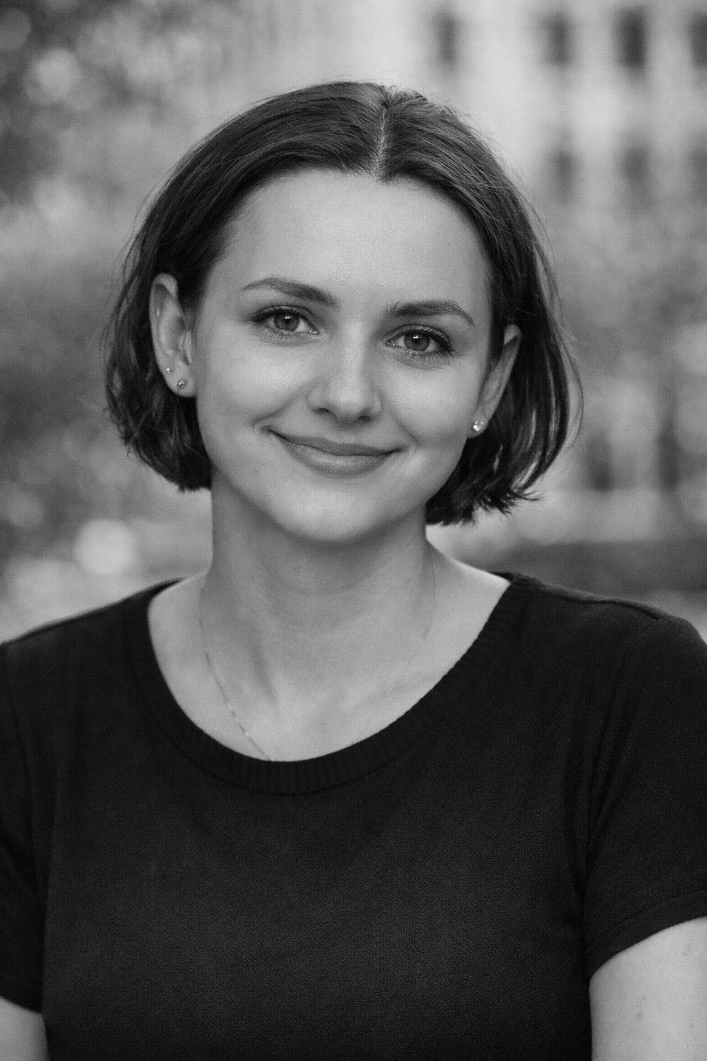 Schwarz-weiß-Porträt einer lächelnden jungen Frau mit kurzen dunklen Haaren, getragenem schwarzen Oberteil und kleinen Ohrringen, draußen mit unscharfem Hintergrund.
