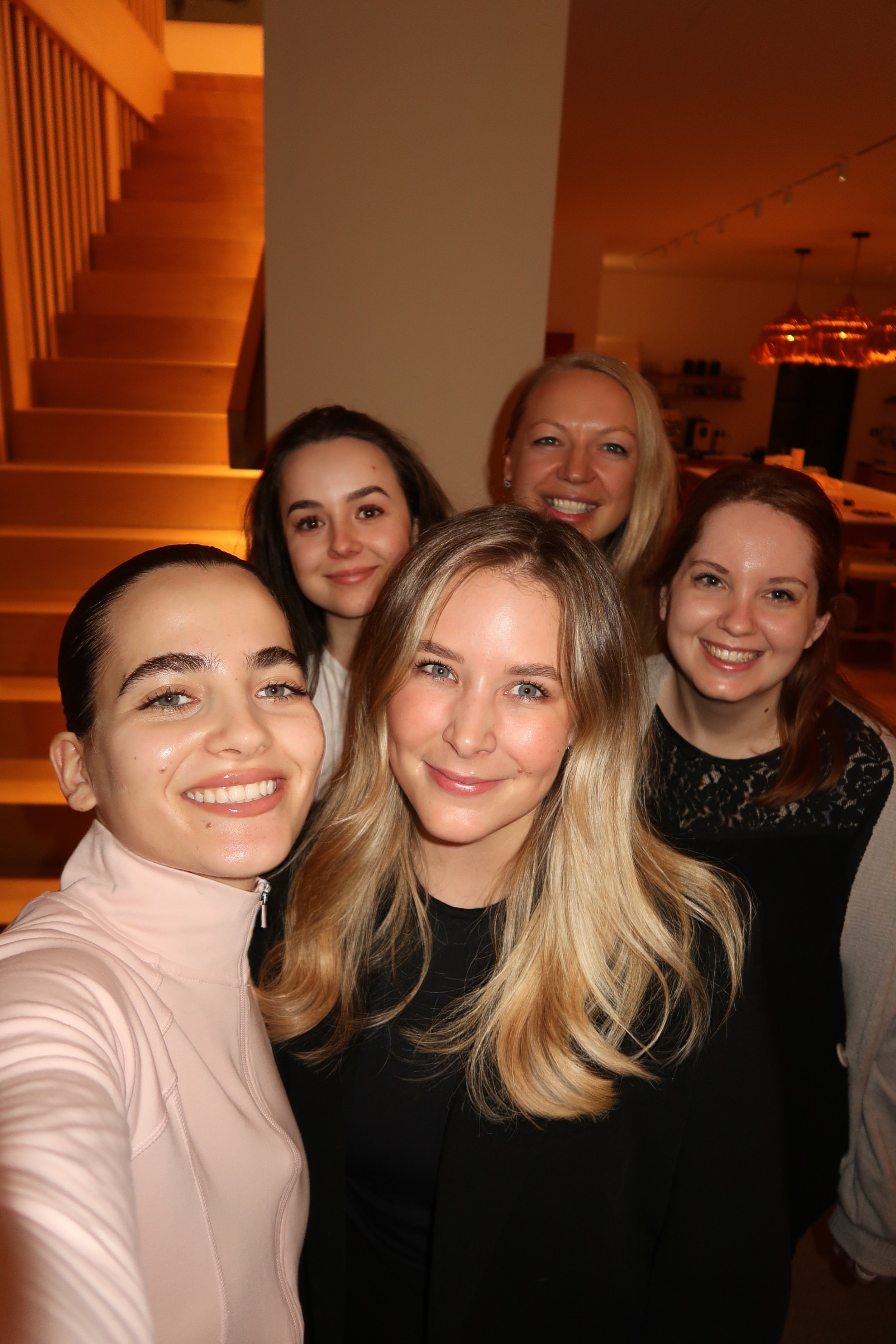 Sechs Frauen, die selfie machen und smiling posieren in einem modernen, warm beleuchteten Raum mit Holztreppe im Hintergrund.