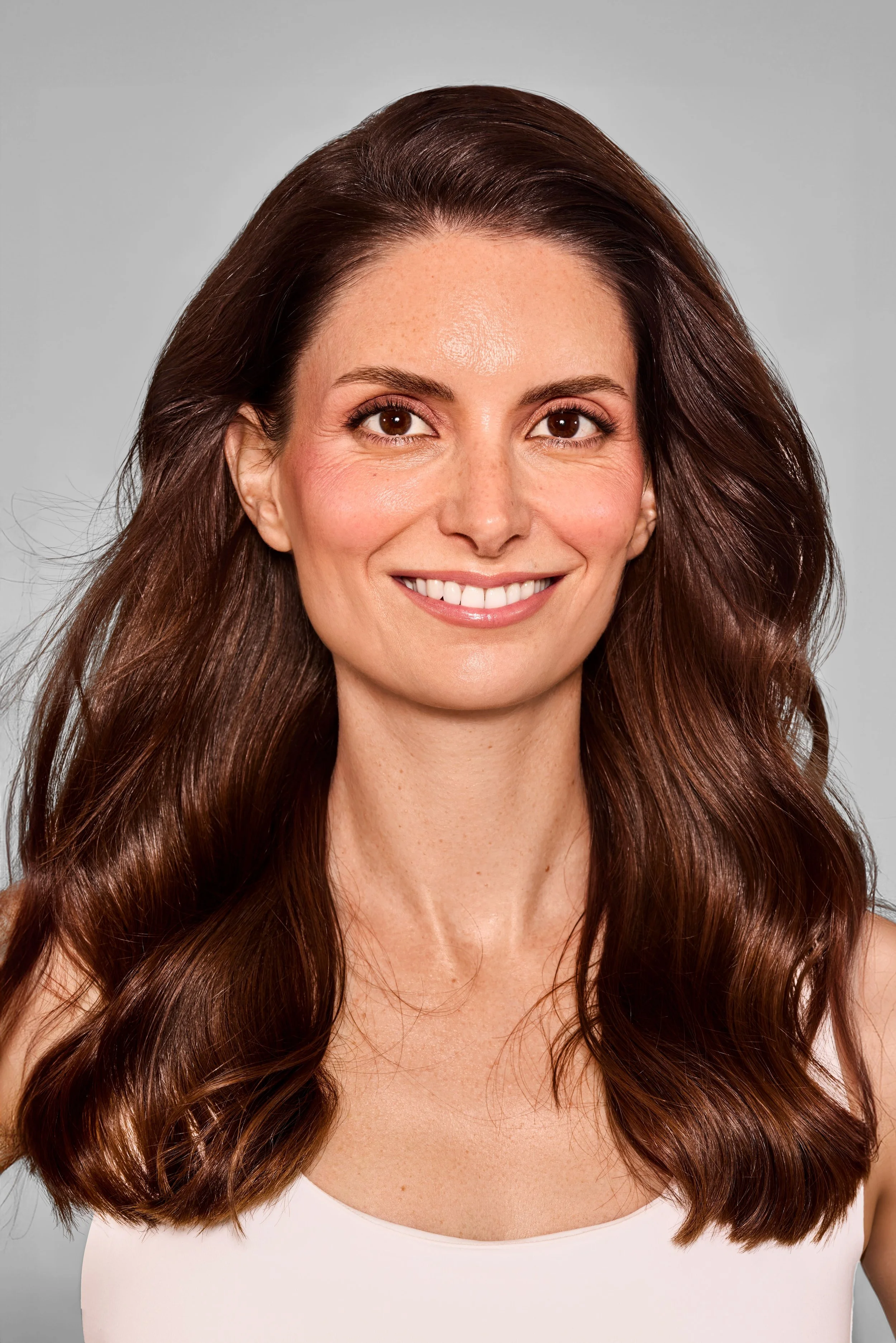 Porträt einer lächelnden Frau mit langen, braunen, lockigen Haaren vor hellem grauem Hintergrund.