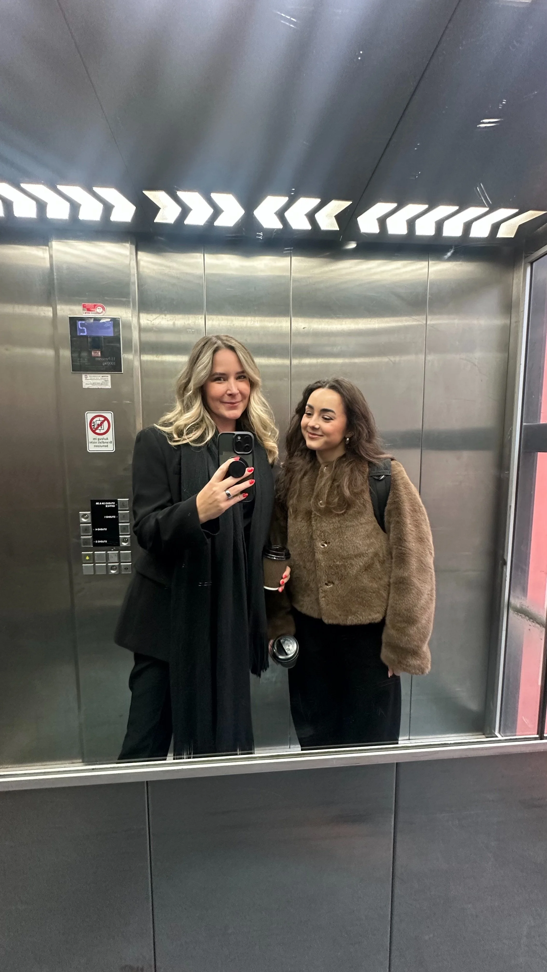 Zwei Frauen nehmen ein Selfie in einem Aufzug mit spiegelndem Wand und Decke. Eine Frau trägt schwarze Kleidung und hält eine Tasse, die andere trägt braune Jacke.