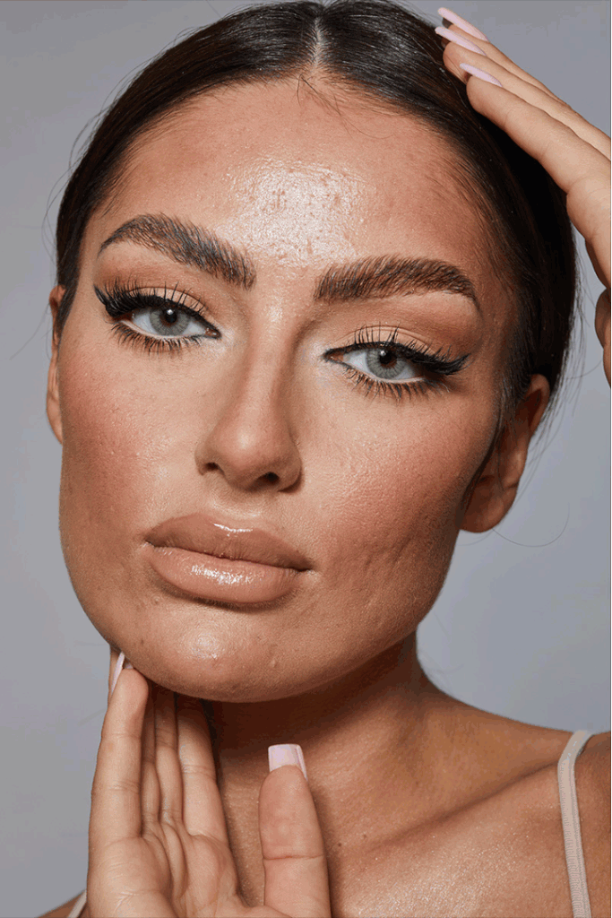 Nahaufnahme eines Models mit glattem, nassem Make-up, strahlender Haut und langen Wimpern, die ihre Augen betonen, während sie ihre Hand an den Kopf legt.