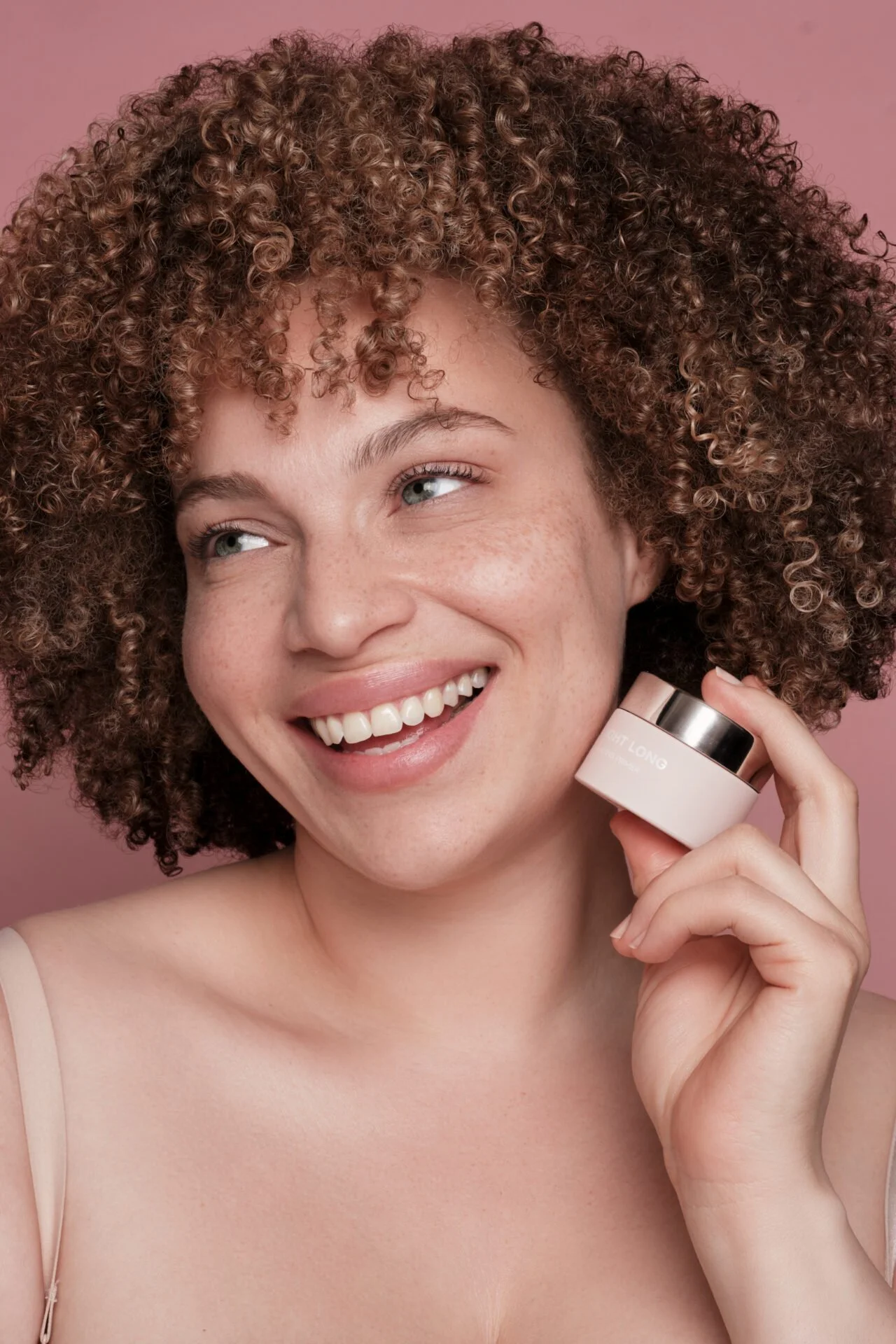 Lächelnde junge Frau mit lockigem Haar hält ein Kosmetikprodukt, vermutlich eine Creme, vor ihr Gesicht. Sie trägt ein hauchzartes Oberteil vor einem rosa Hintergrund.