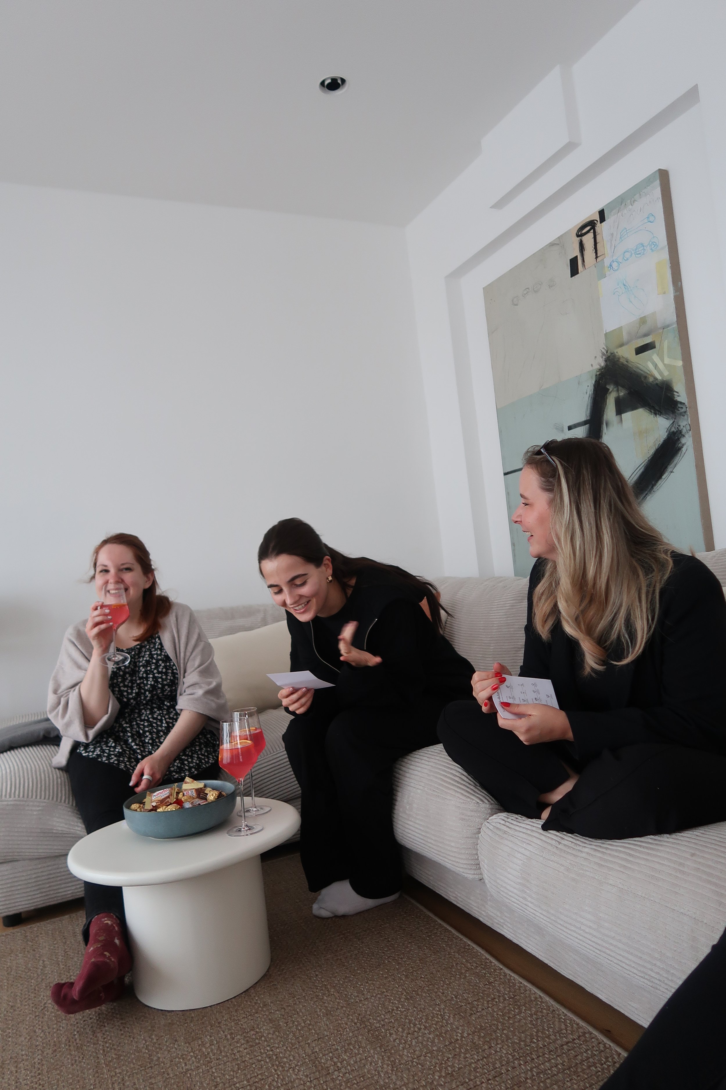 Drei Frauen sitzen auf einer Couch, lachen und unterhalten sich. Es gibt ein Glas mit rotem Getränk, eine Schüssel mit Süßigkeiten auf einem kleinen weißen Tisch vor ihnen. Die Dekoration ist modern, mit einem großen abstrakten Gemälde an der Wand.