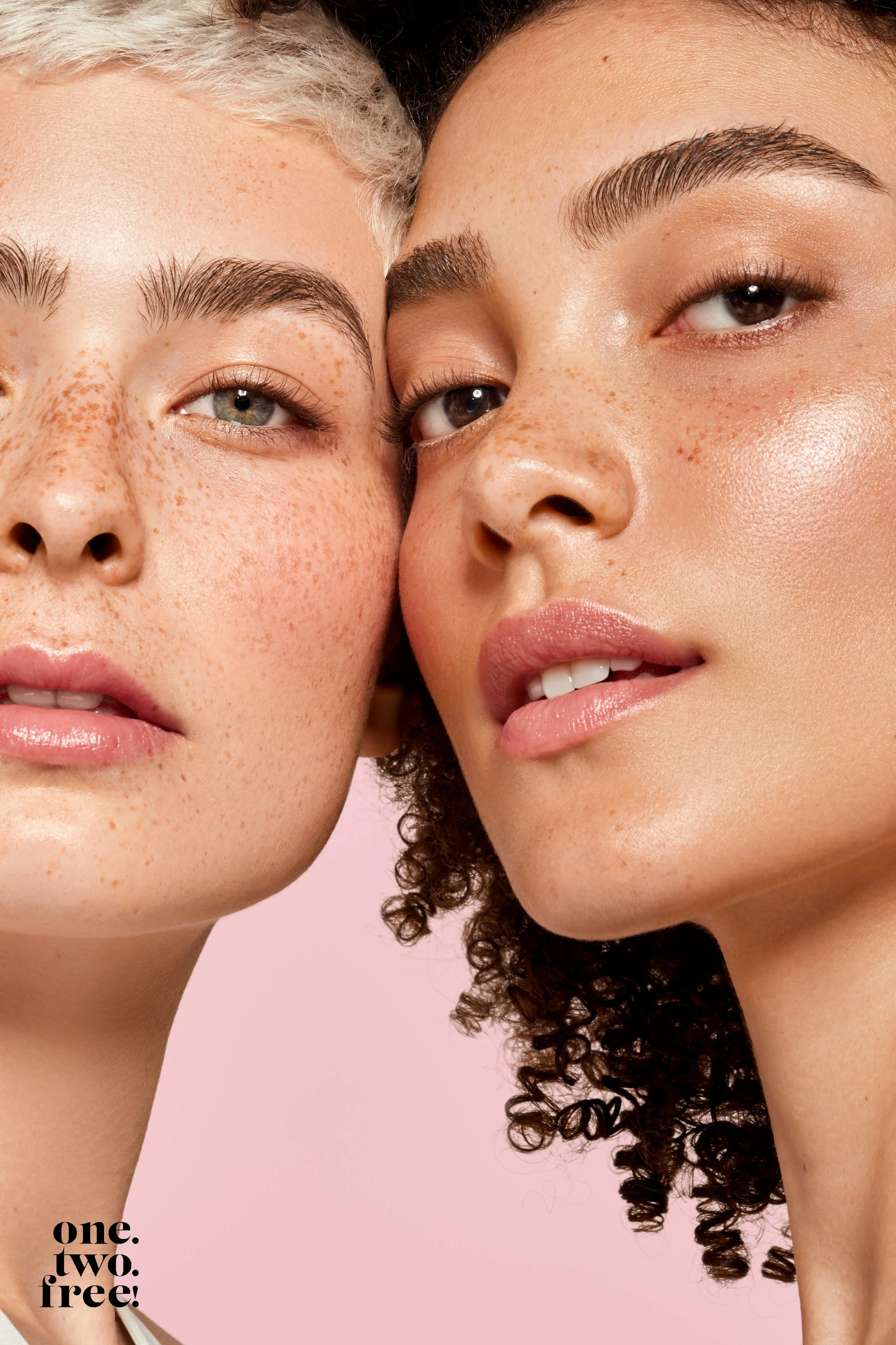 Nahaufnahme von zwei Frauen mit natürlicher Haut, die sich nahekommen, vor einem hellrosa Hintergrund. Text in der unteren linken Ecke: "one. two. free!"