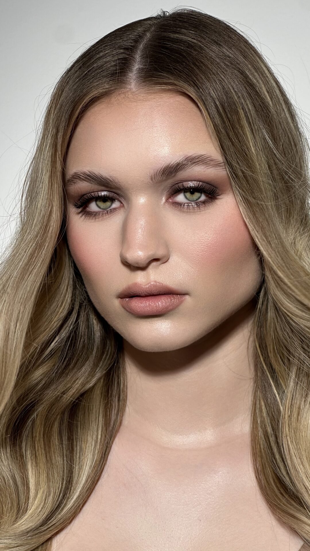 Nahaufnahme eines jungen Mädchens mit langen, gewellten, braunen Haaren und grünen Augen, leichter Make-up und neutralem Gesichtsausdruck.