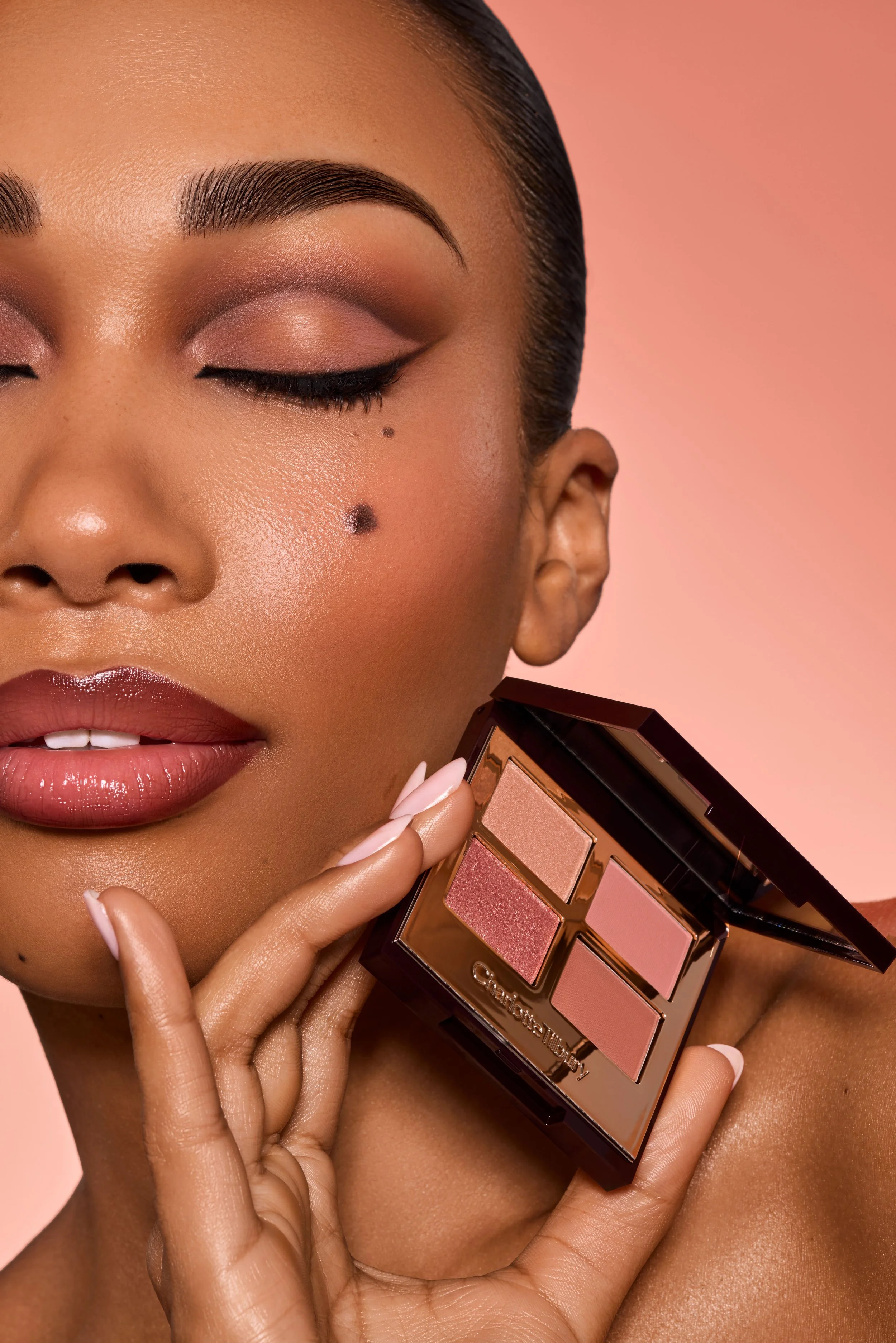 Nahaufnahme eines Models mit geschminktem Gesicht, das einen Beauty-Pinsel hält, der ein Make-up-Palettenprodukt mit fünf Lidschatten-Farben zeigt, vor einem rosafarbenen Hintergrund.