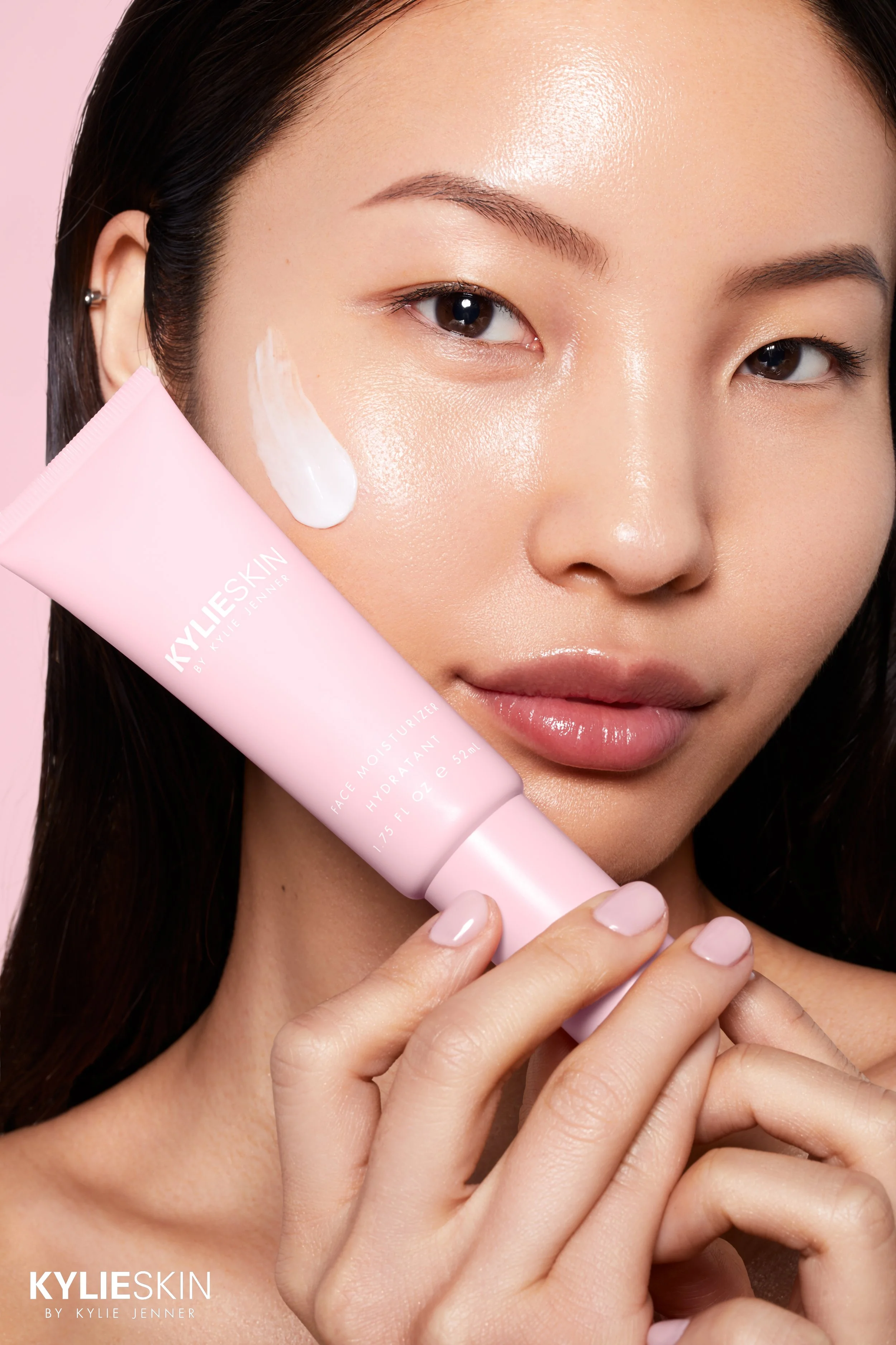 Nahaufnahme einer Frau mit makelloser Haut, die ein rosa Tubenprodukt von KYLIESKIN hält, das auf ihre Wange aufgetragen wurde.