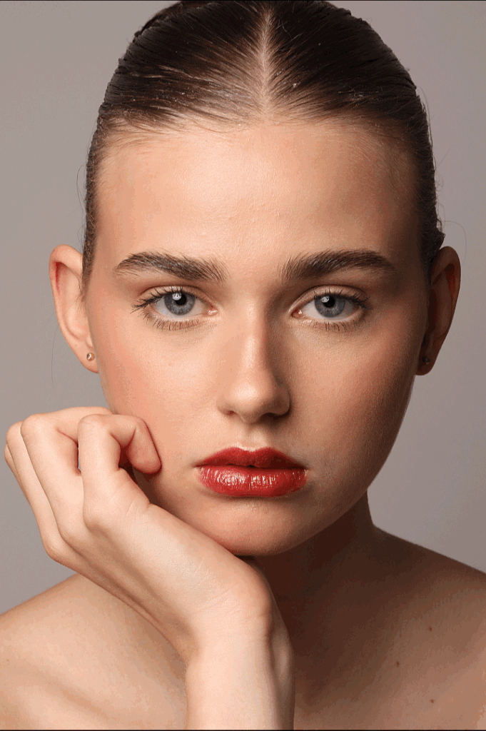 Nahaufnahme eines jungen Mädchens mit blauen Augen, glatter Haut, roten Lippen und dunklem, zurückgegelt Haar, das an einem grauen Hintergrund lehnt.