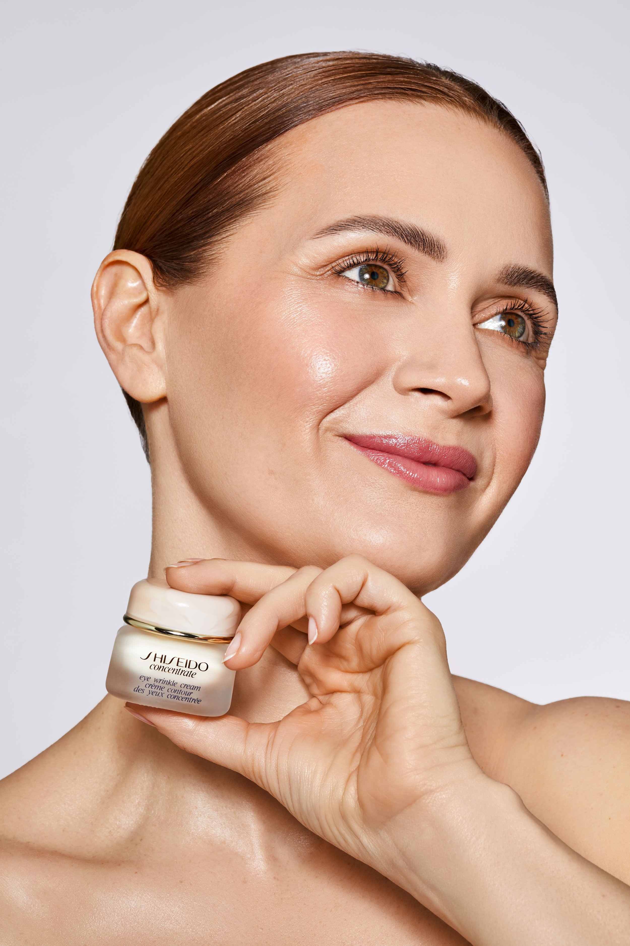 Naheaufnahme eines lachenden Gesichts einer Frau mit glatter Haut, die eine Creme hält, die als 'Shiseido Concentrate Eye Wrinkle Cream' gekennzeichnet ist.