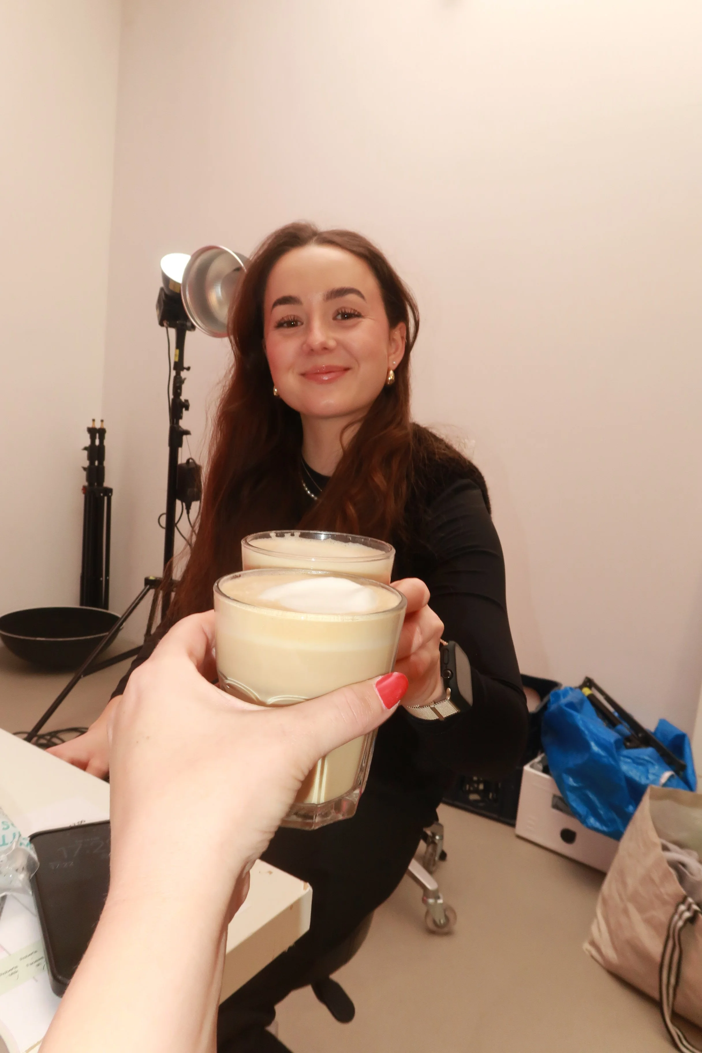 Zwei Menschen stoßen mit Getränken an, eine davon ist eine lachende Frau mit braunen Haaren und schwarzen Kleidung, im Hintergrund sind Fotografieausrüstung und ein Raum mit Gegenständen.