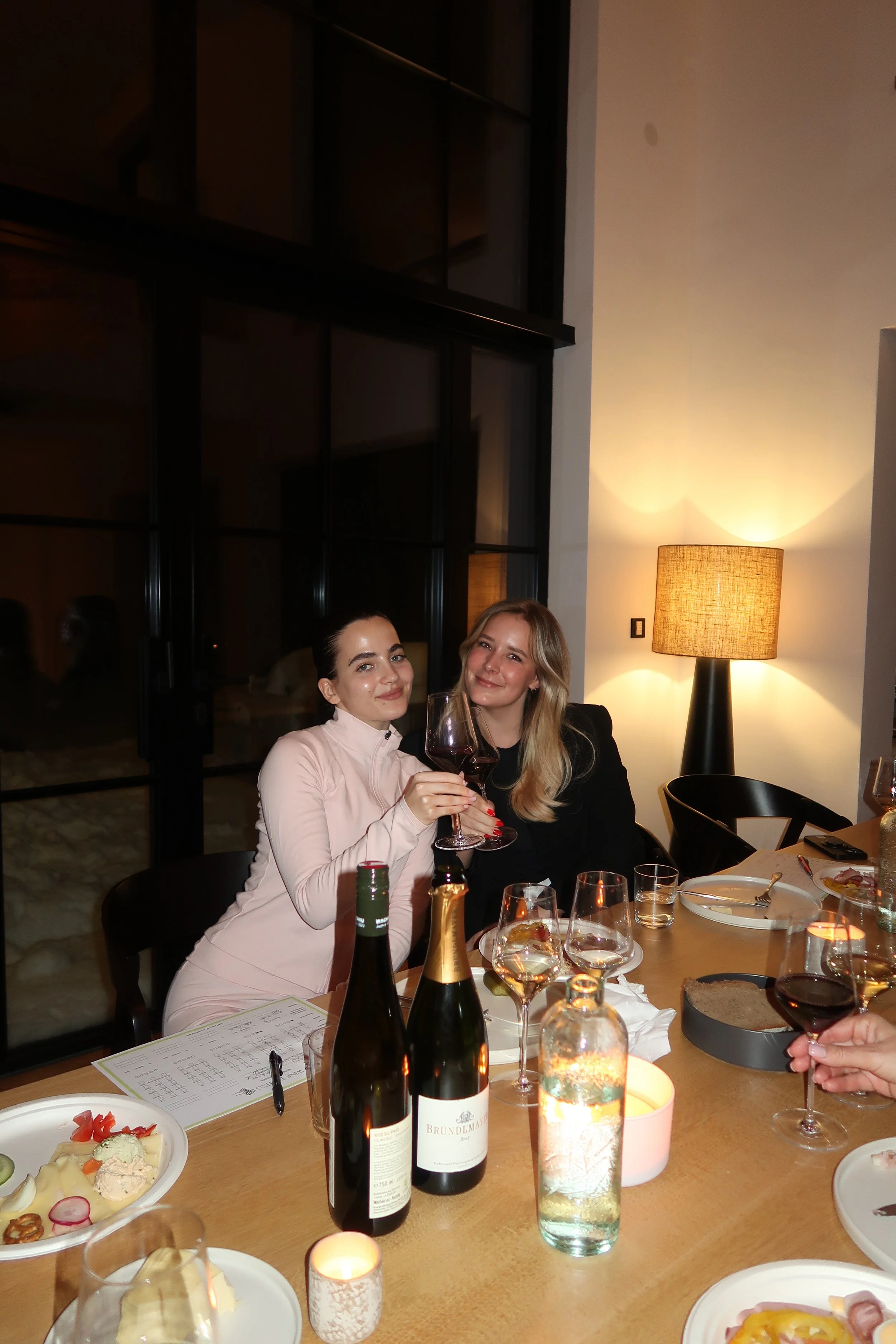 Zwei Frauen sitzen an einem Esstisch, halten rote Weingläser und lachen. Der Tisch ist mit Wein, Wasser, Kerzen und Tellern gedeckt. Es ist dunkel draußen, im Hintergrund ein großes Fenster und eine Stehlampe