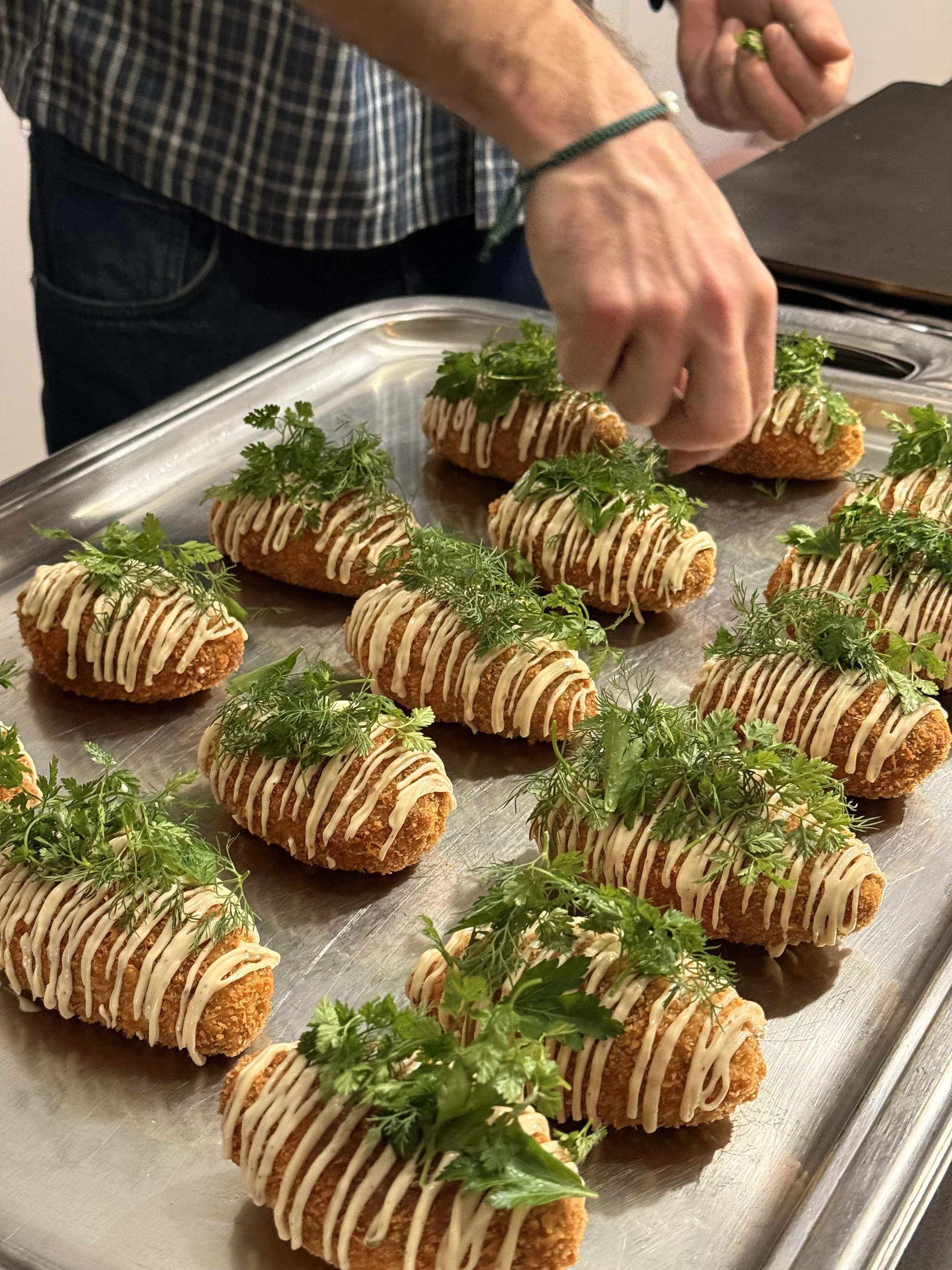 Des croquettes frites garnies de persil et de sauce blanche, disposées sur une plaque en métal, avec une personne décorant les plats.