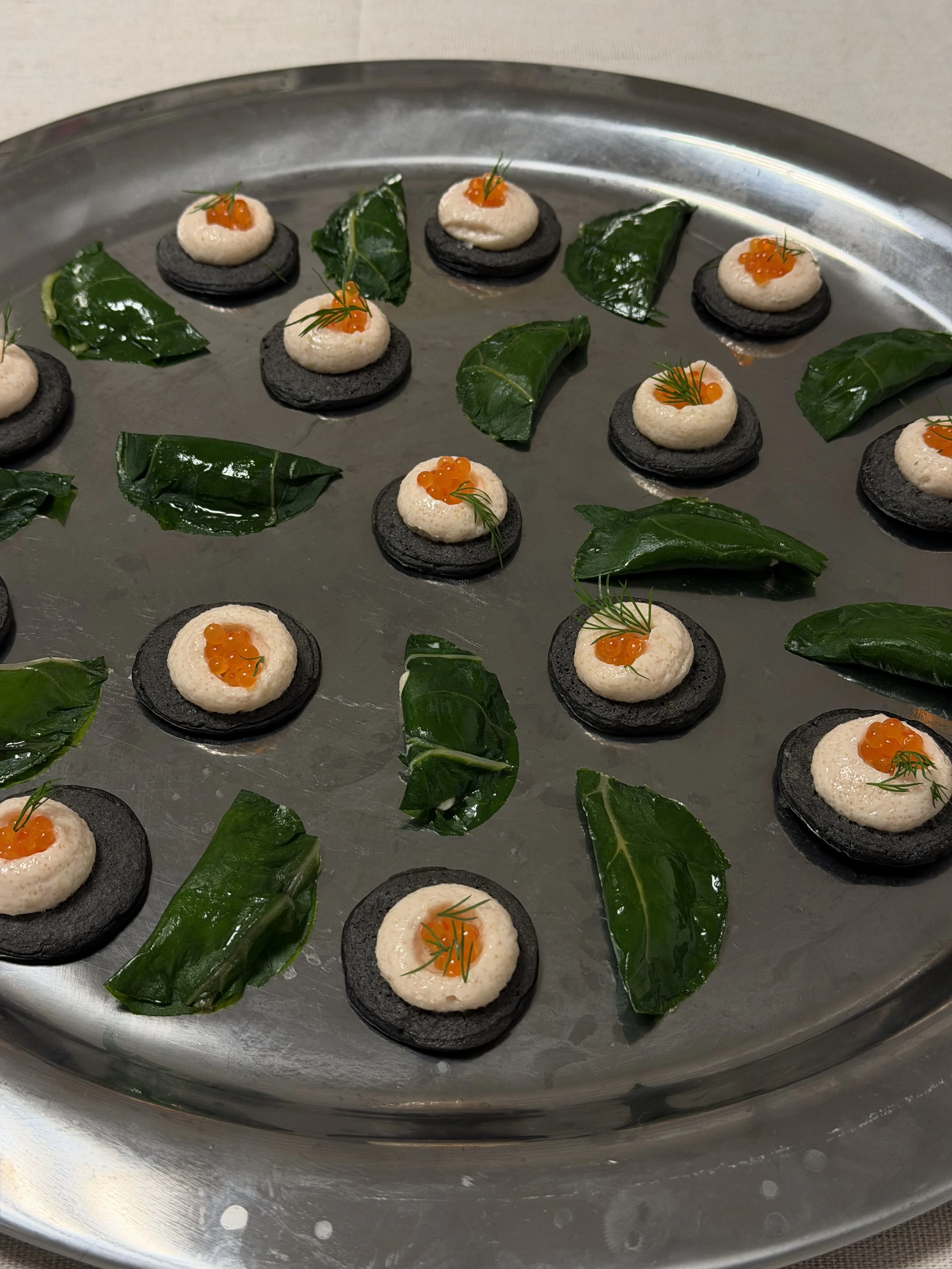 Amuse-bouche avec une base noire, garniture blanche, œufs de saumon orange et une petite branche d'aneth, accompagnés de feuilles de salade vert foncé.