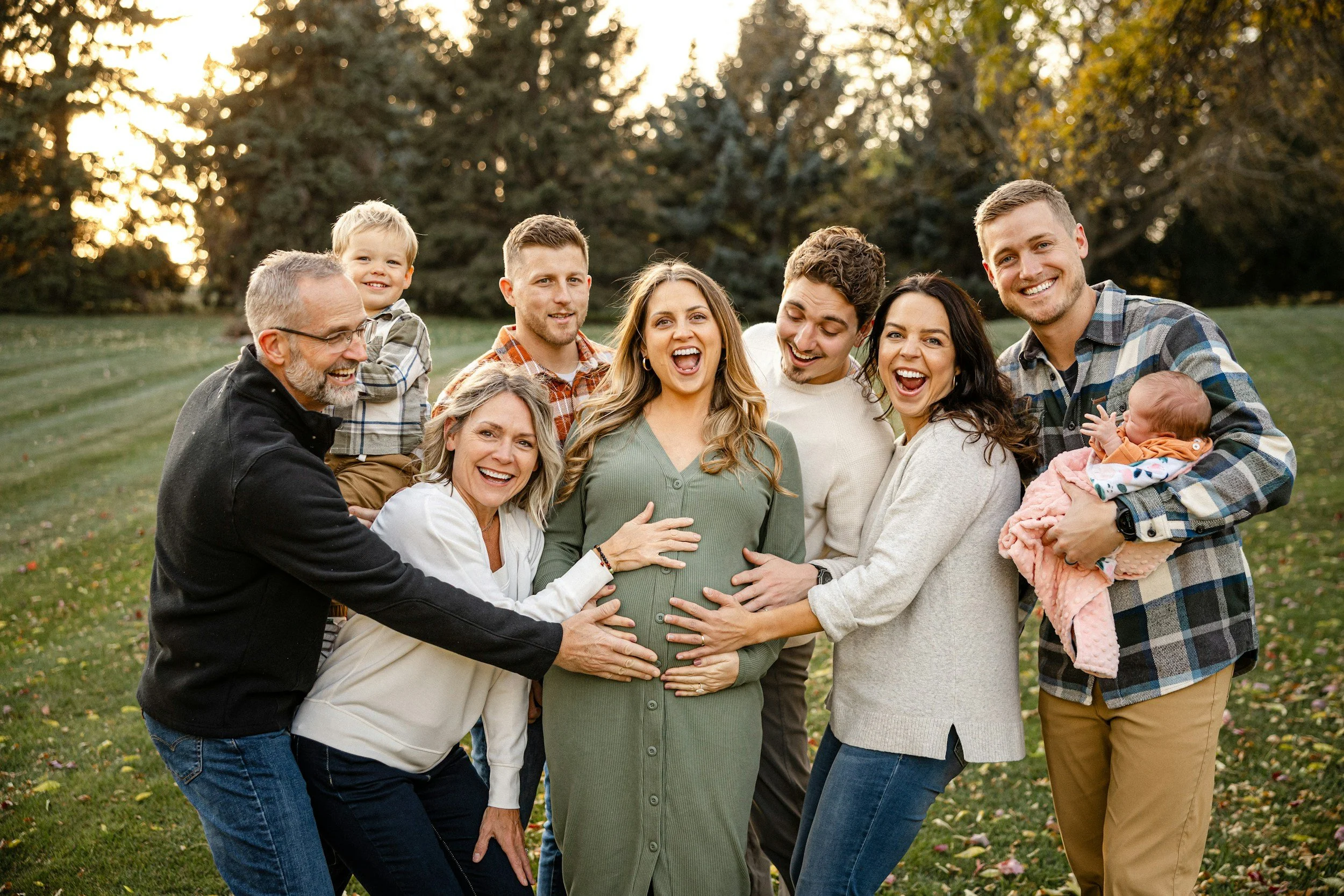 A large family group standing outdoors in a park during autumn, celebrating a pregnant woman in the center with friends and family smiling and touching her belly.