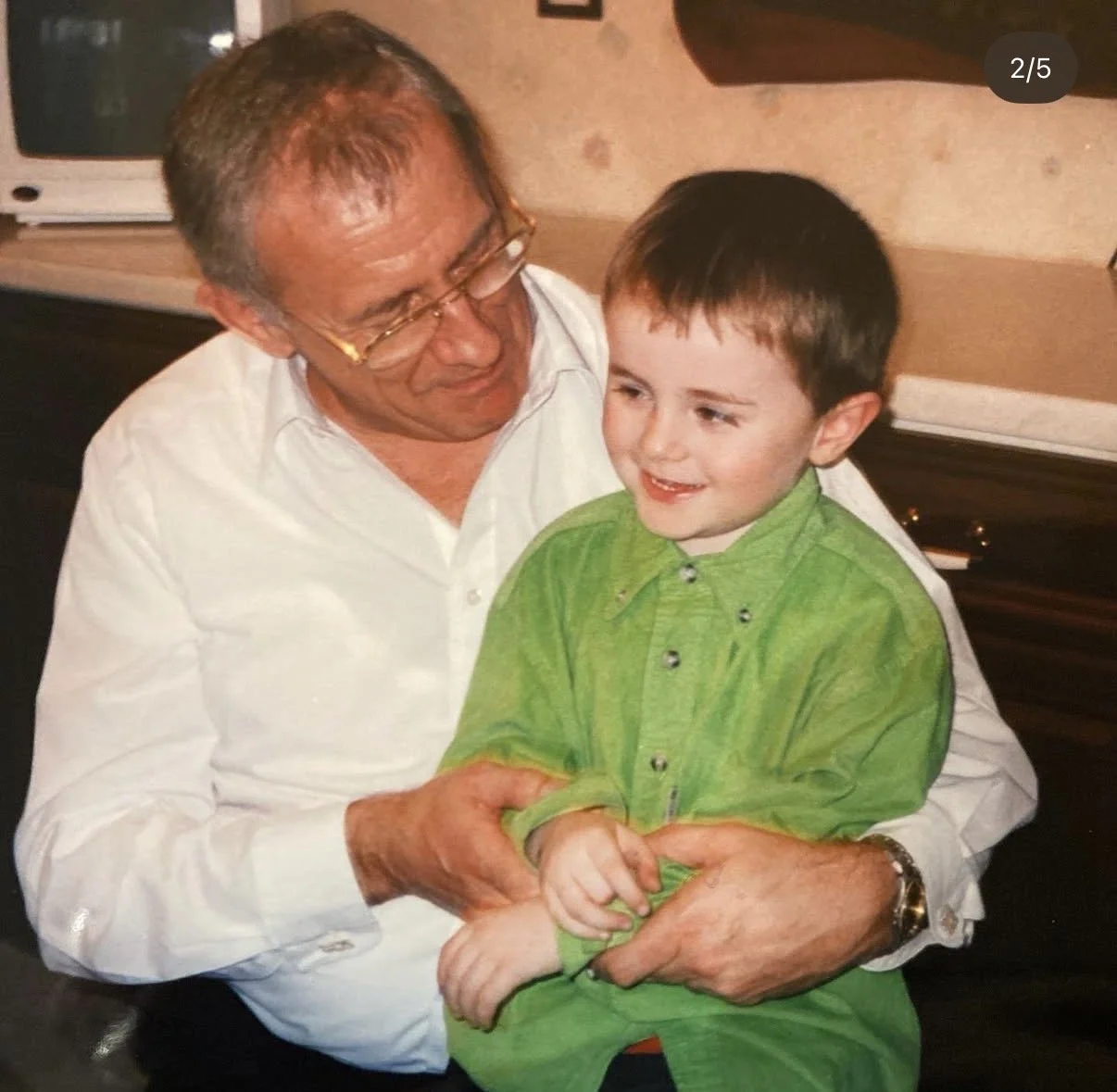 An older man with glasses and a white shirt holding a young boy in a green shirt, both smiling and playing together inside a room.