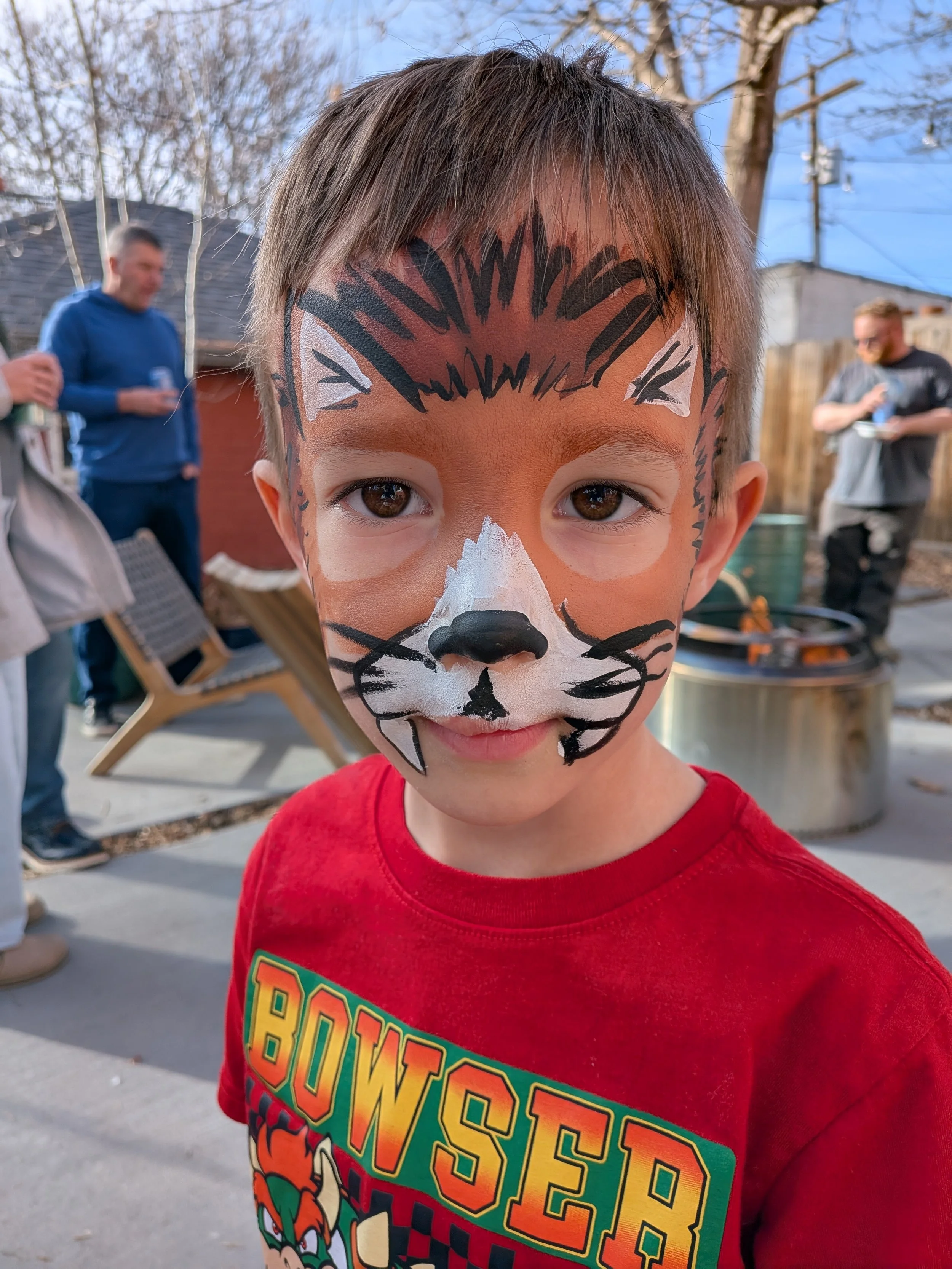 A young boy has face paint resembling a tiger, with orange, white, and black colors, and is wearing a red T-shirt with 