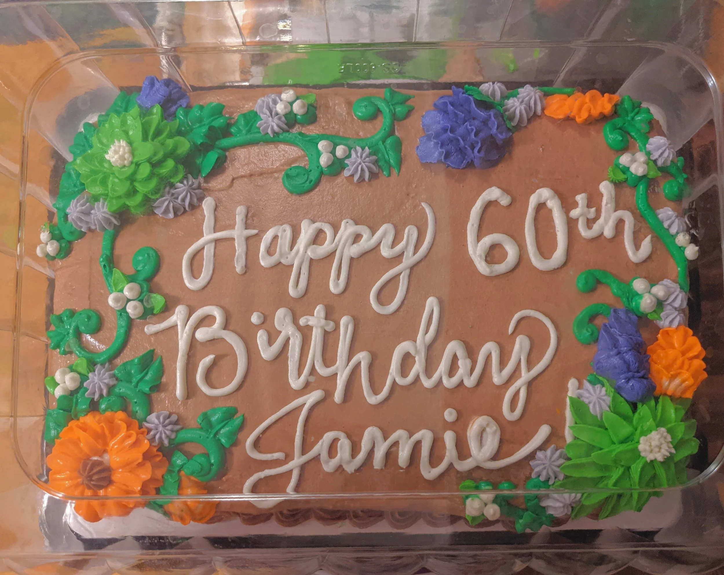 A rectangular birthday cake with brown frosting, decorated with colorful piped flowers and green leaves, reads "Happy 60th Birthday Jamie" in white icing.