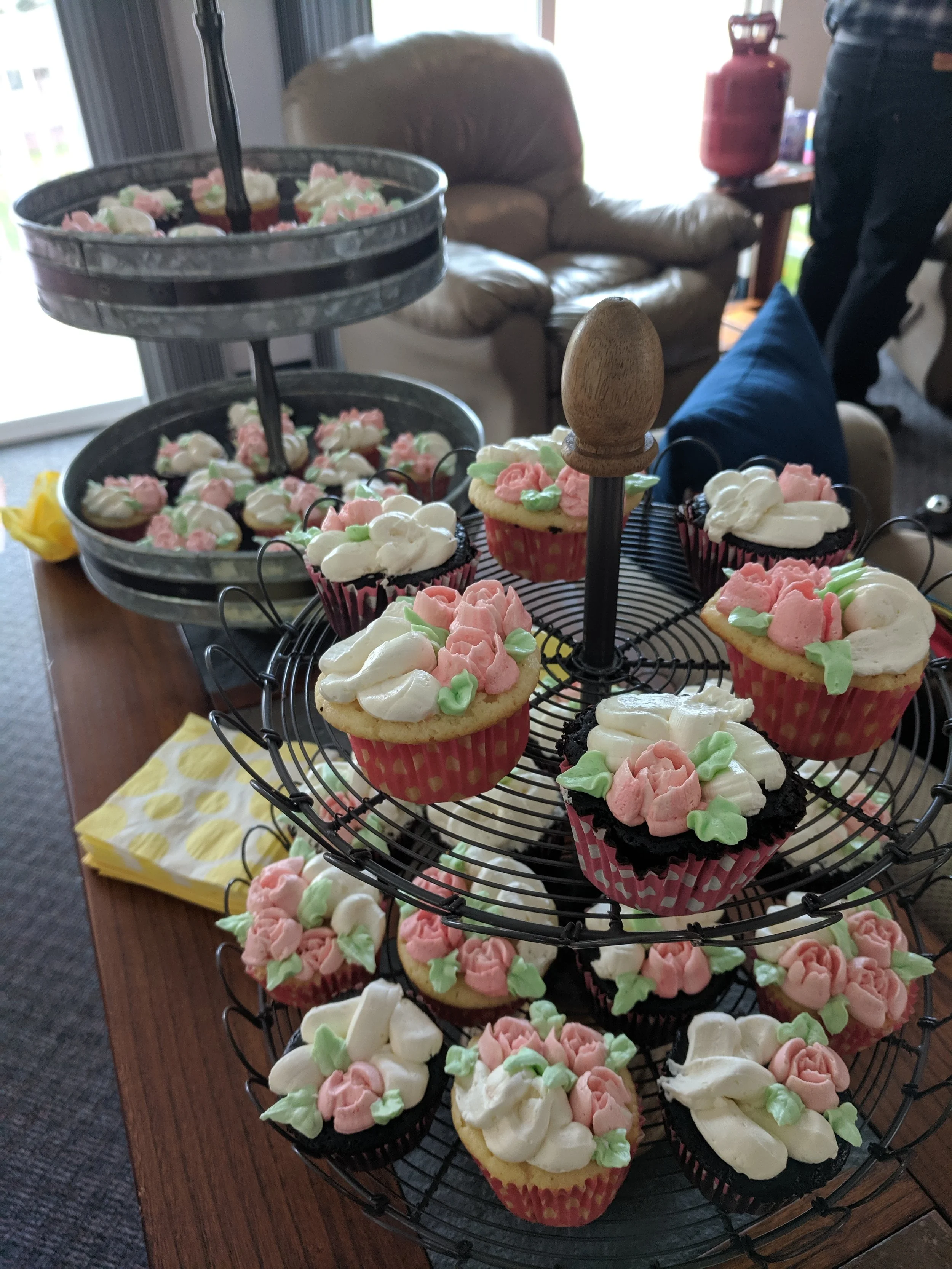Cupcakes decorated with pink roses and green leaves placed on a tiered metal serving tray on a wooden table.