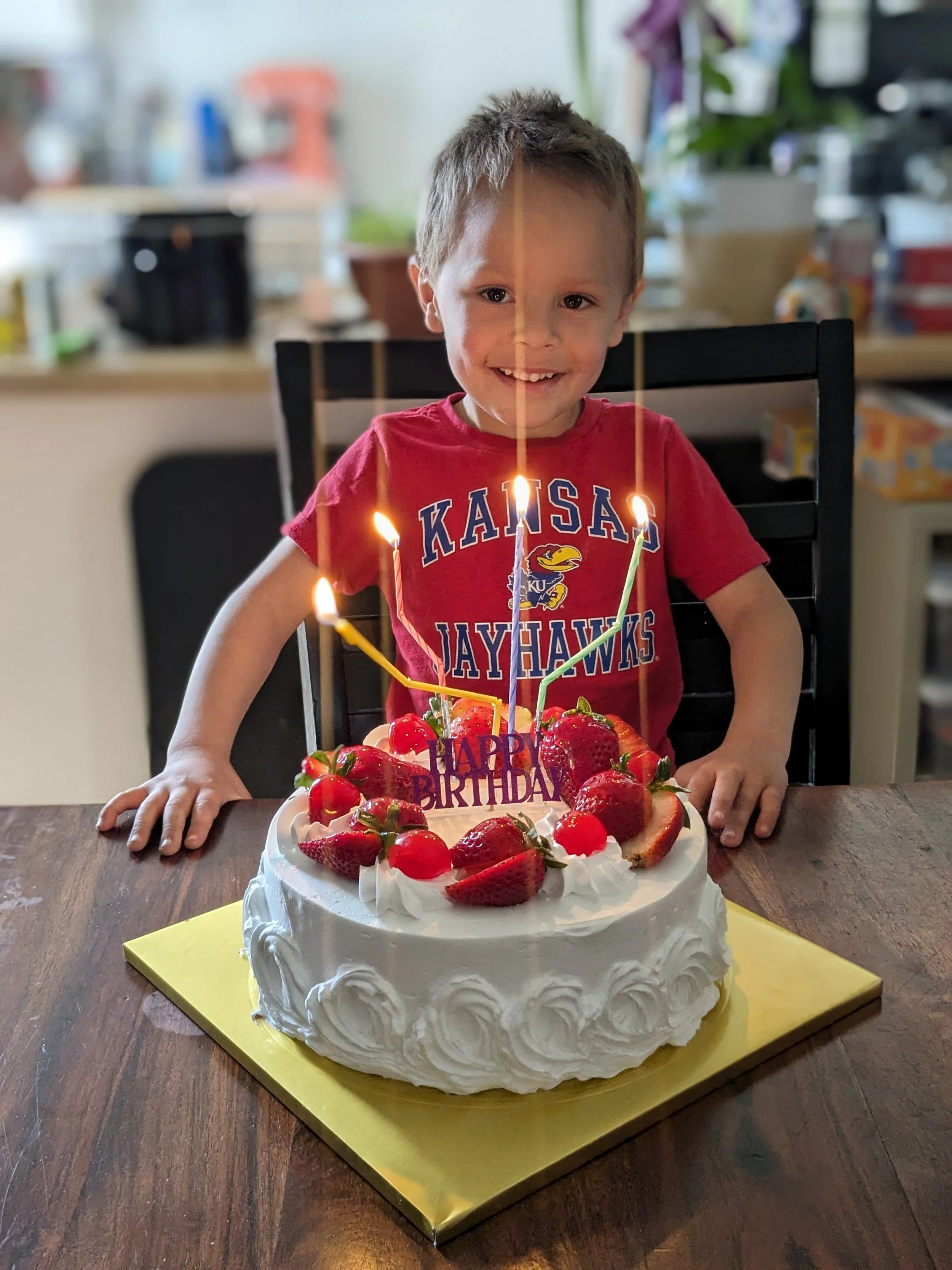 A young boy sitting at a table with a birthday cake decorated with strawberries and candles, wearing a red Kansas Jayhawks t-shirt, smiling.