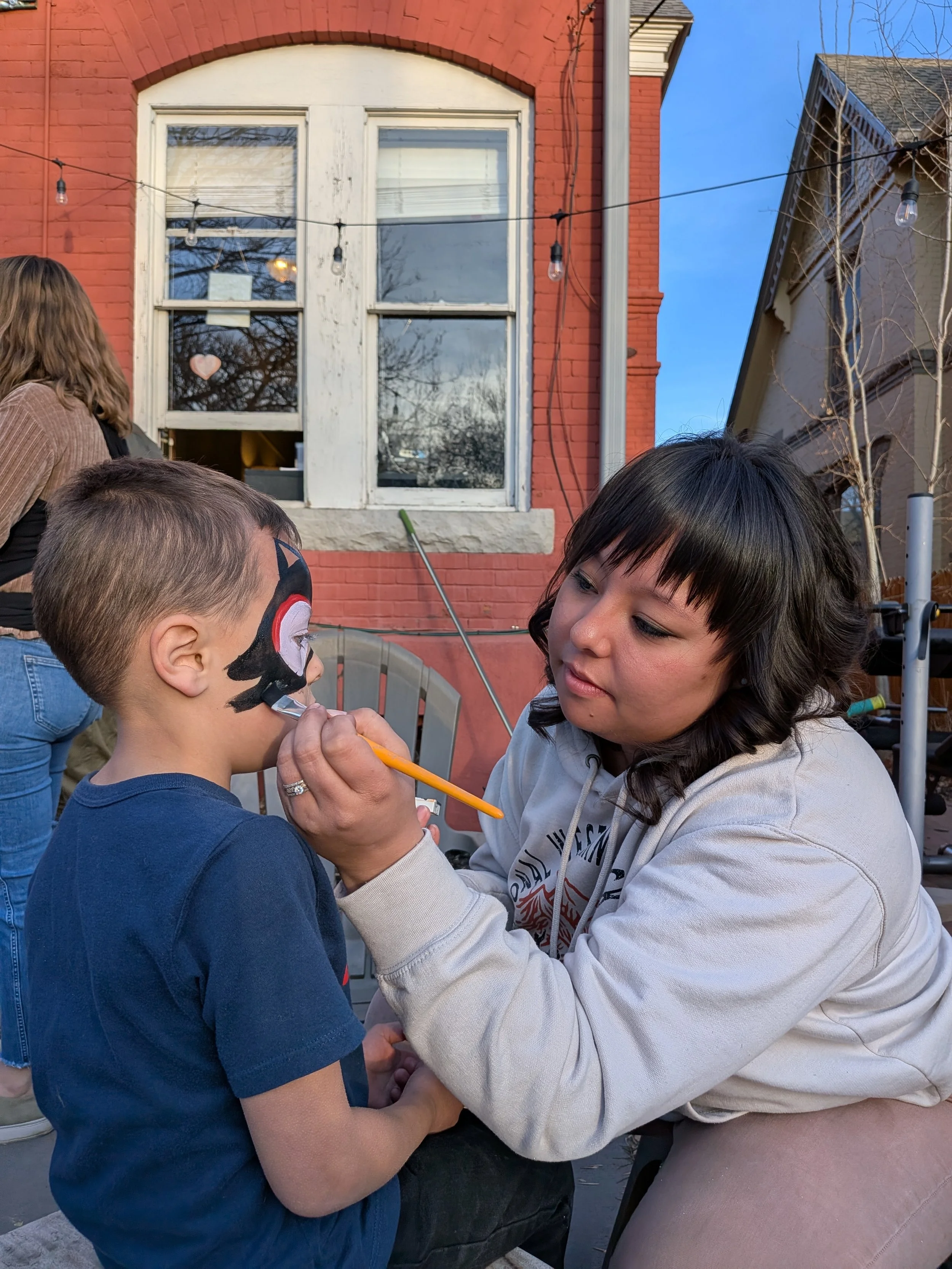 A young girl with face paint being carefully applied by an adult woman outdoors. The face paint resembles a character with black, red, and white colors. The scene takes place in front of a red brick building with large windows, string lights, and a c
