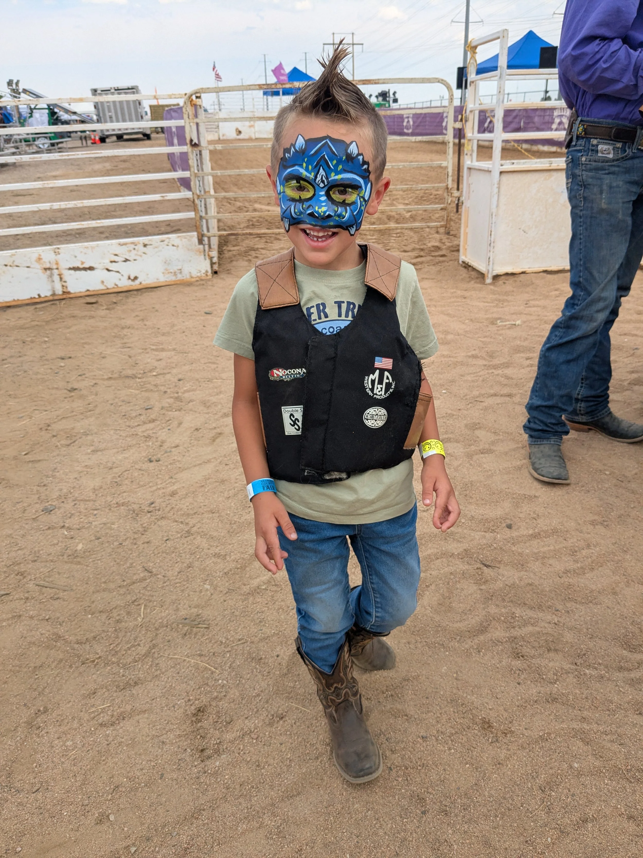 A smiling young boy with a painted blue and green dragon mask on his face, dressed in cowboy attire with a vest, jeans, and cowboy boots at a rodeo or fairground.