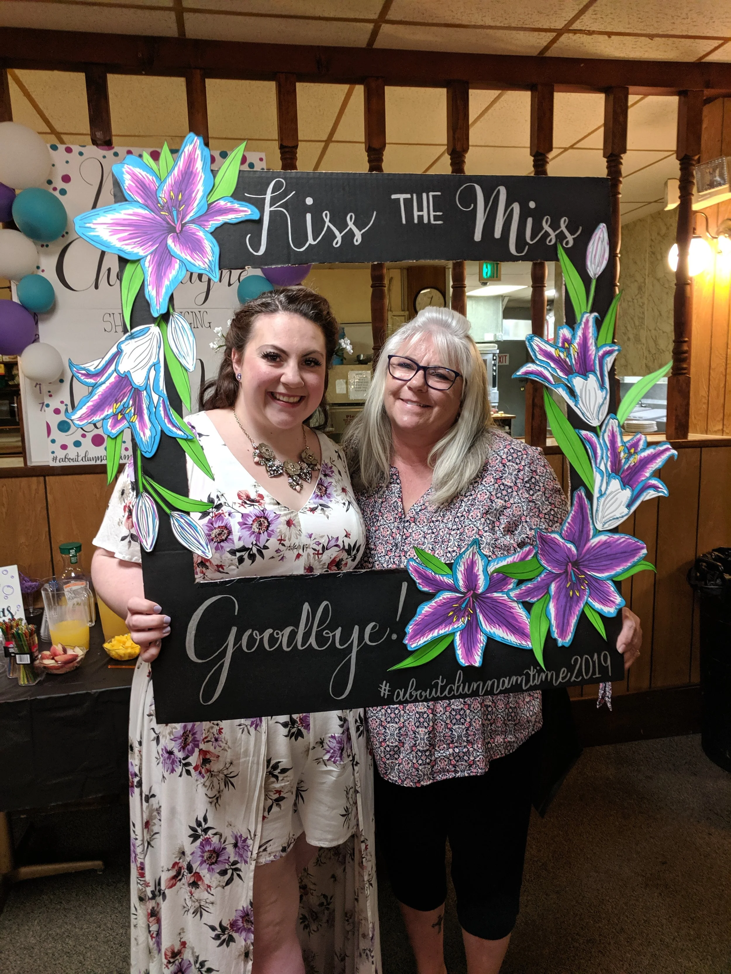 Two women smiling and holding a black sign with purple lilies and white text, at a farewell event. The sign reads 'Kiss the Miss Goodbye! #aboutclammamtime2019'.