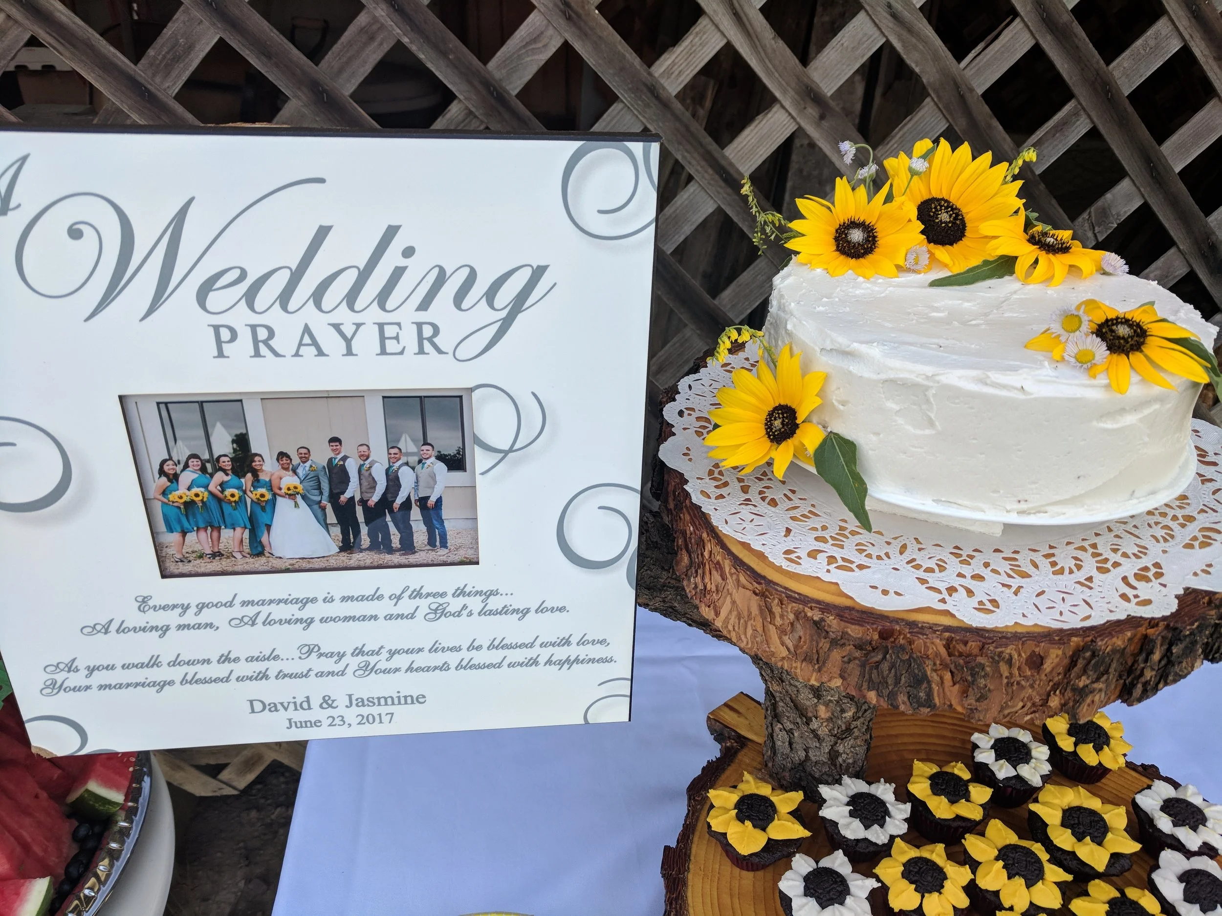 Wedding table with a sign saying 'Wedding Prayer' and a photo of a wedding party. The table has a decorated cake with yellow sunflowers and small white daisies, placed on a wooden stand with a doily underneath.