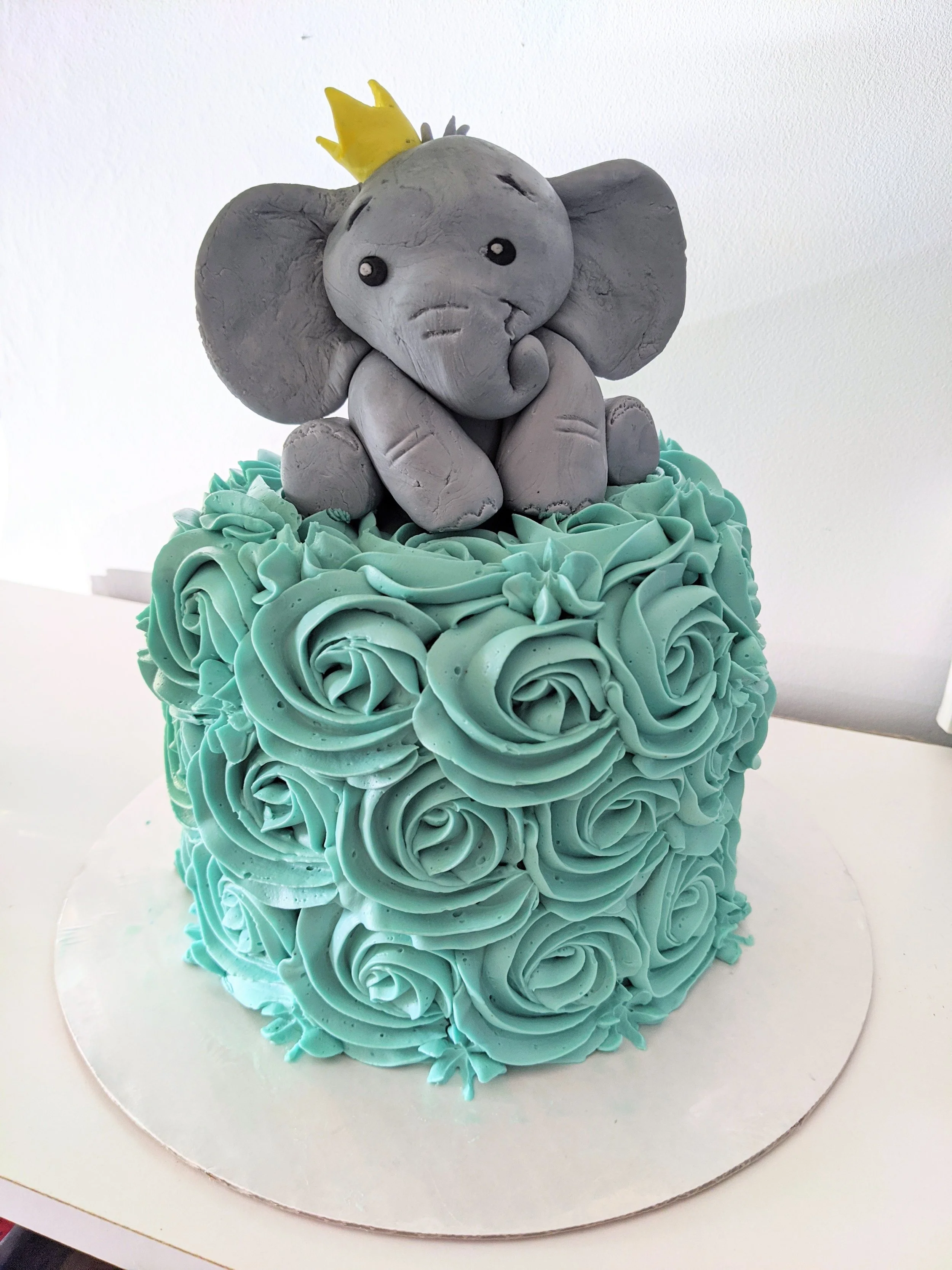 A cake decorated with light blue frosting in rosette patterns and a gray elephant figurine topper wearing a yellow crown.