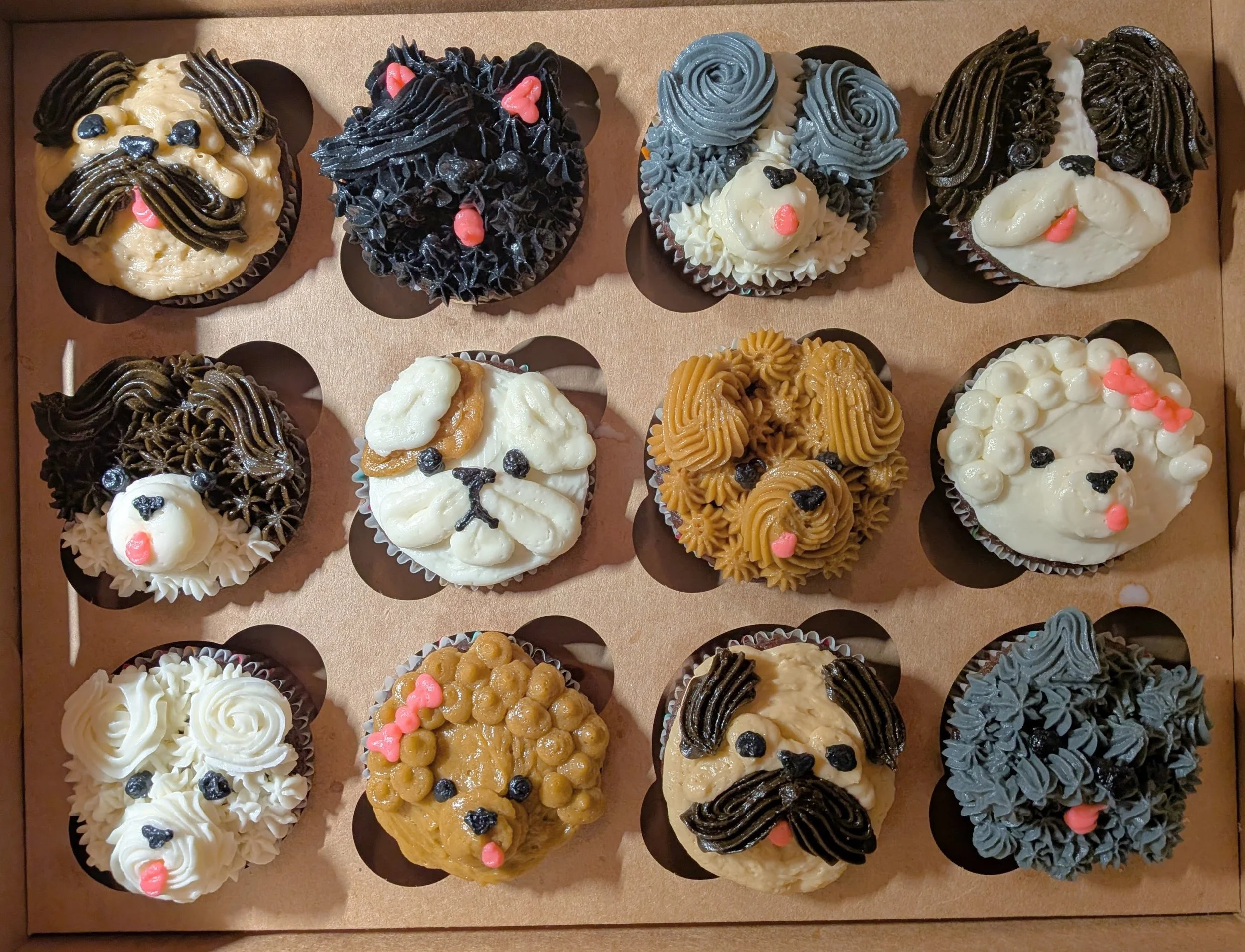 A box of 15 decorated cupcakes with dog faces, each with different frosting designs and dog features like ears, eyes, nose, and tongue.