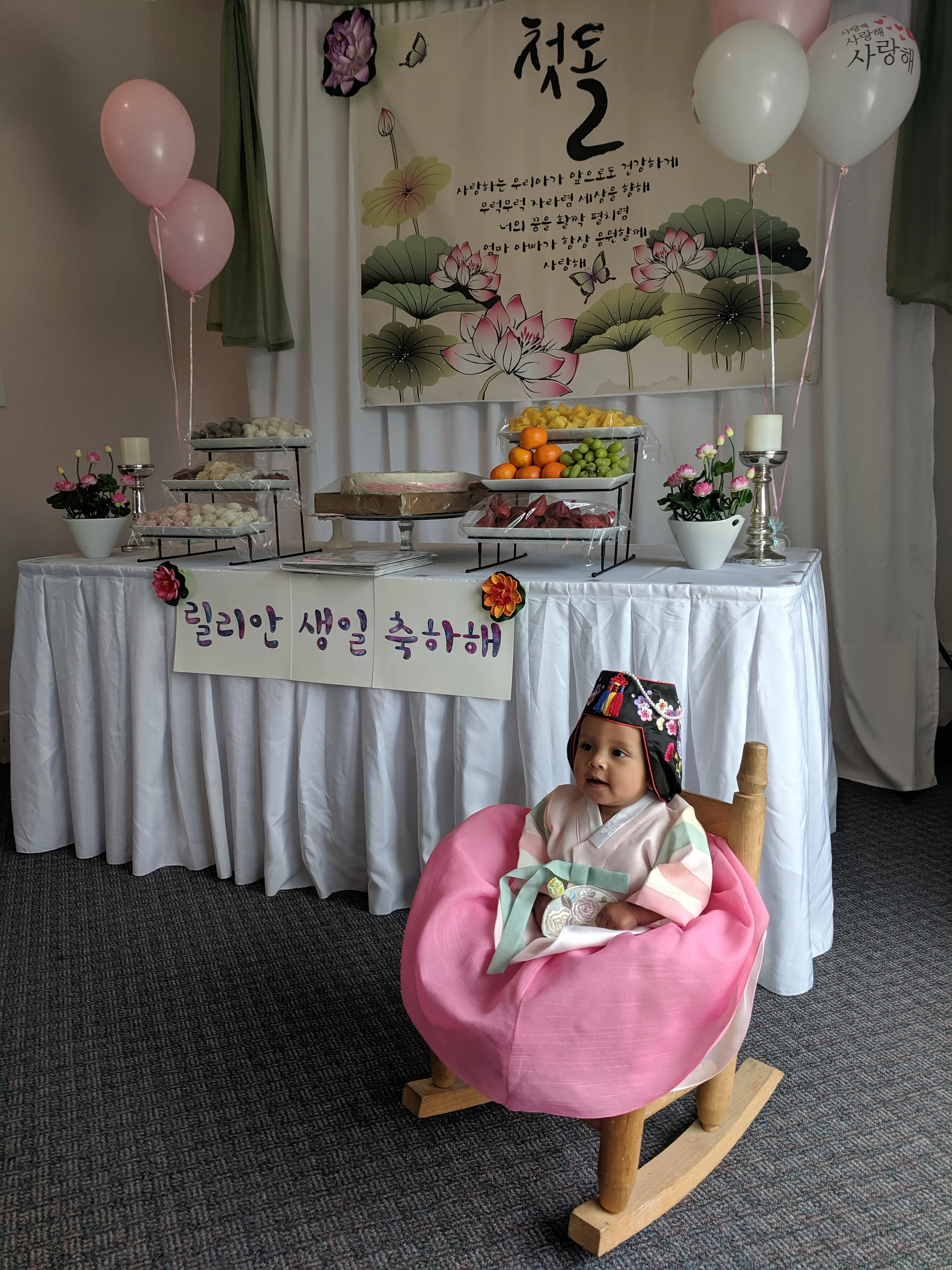 A young child dressed in traditional Korean clothing sitting in a pink cushioned wooden rocking chair in front of a decorated table with food and balloons, celebrating a birthday with a 'Happy Birthday' sign in Korean.