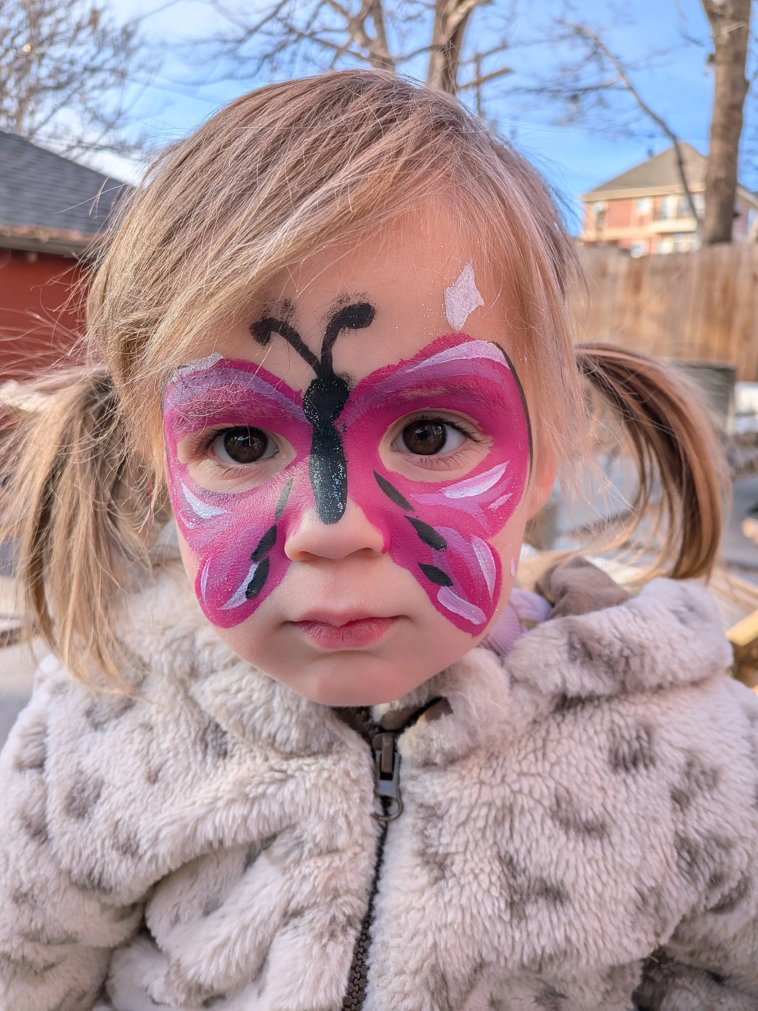 A young girl with face paint of a pink butterfly with black and white details, wearing a beige furry jacket, outdoors on a clear day.