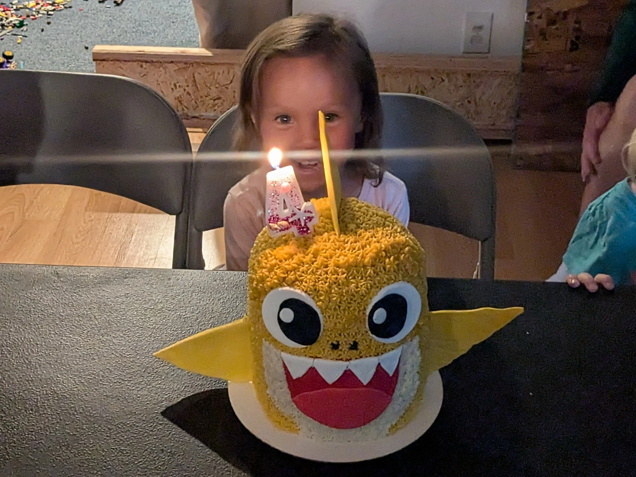 A child with brown hair and a white shirt sitting at a table with a dragon-themed birthday cake with big eyes, yellow ears, and a wide open mouth with white fangs. The cake has a lit birthday candle with the number 4 on it, and a yellow candle. The c