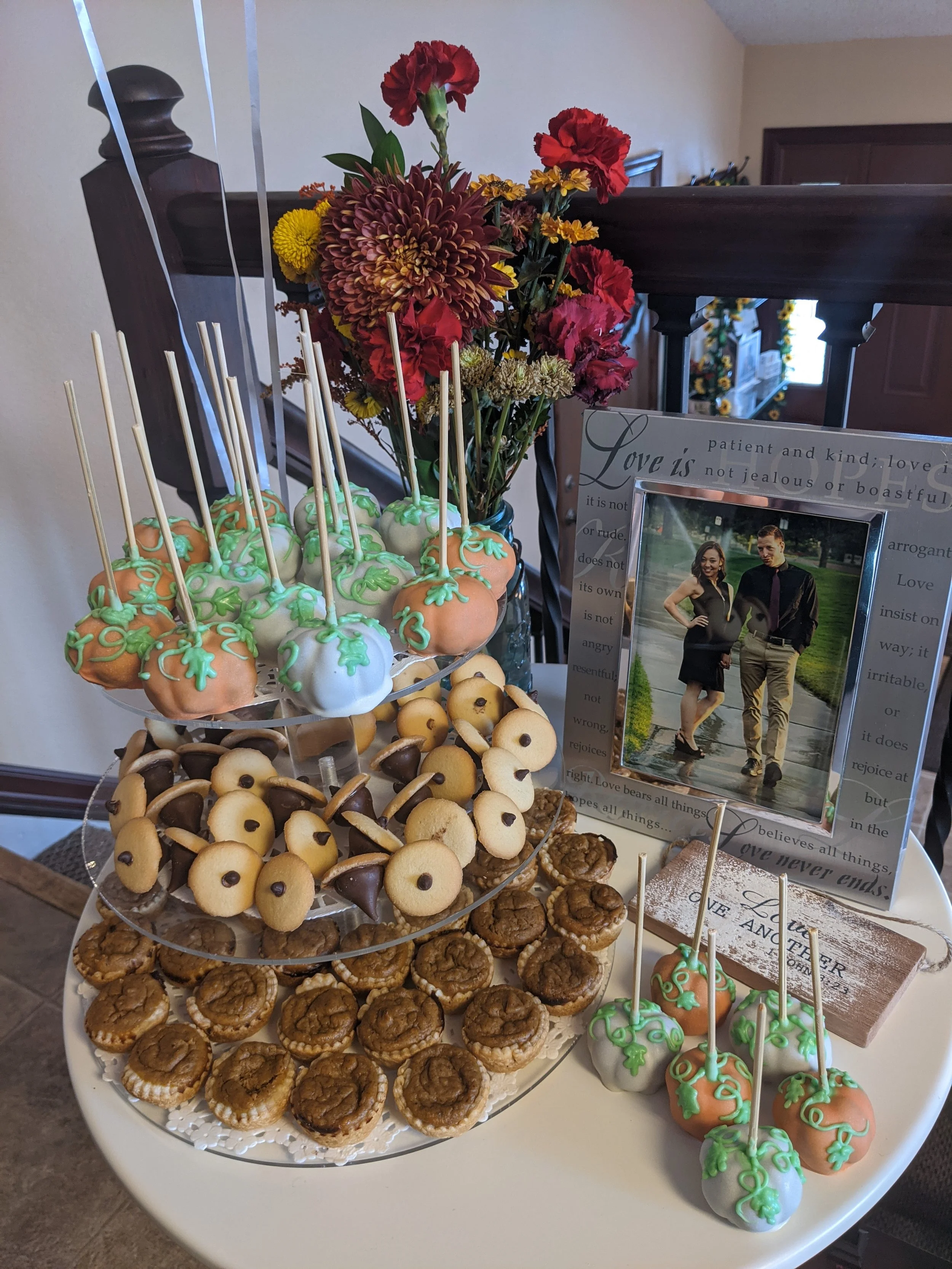 Dessert display with decorated cake pops, cookies, and mini cupcakes on a tiered stand, alongside a flower arrangement, framed photograph, and decorative signs on a round table.