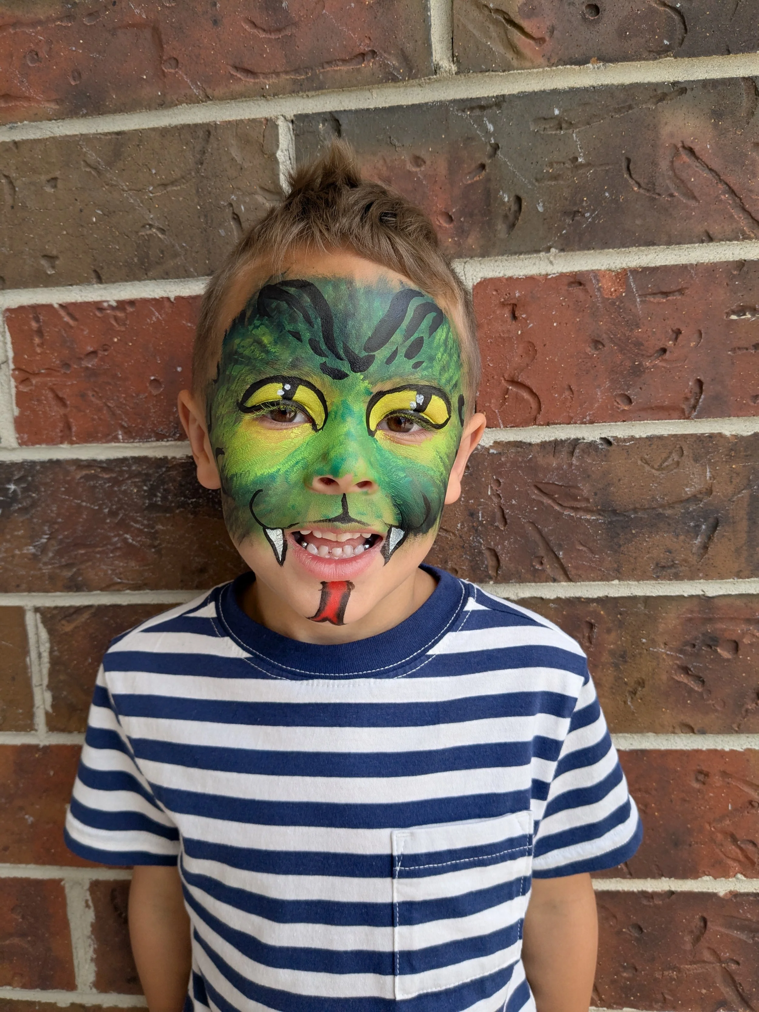 Child with a painted face resembling a green dragon or lizard, smiling and standing against a brick wall.