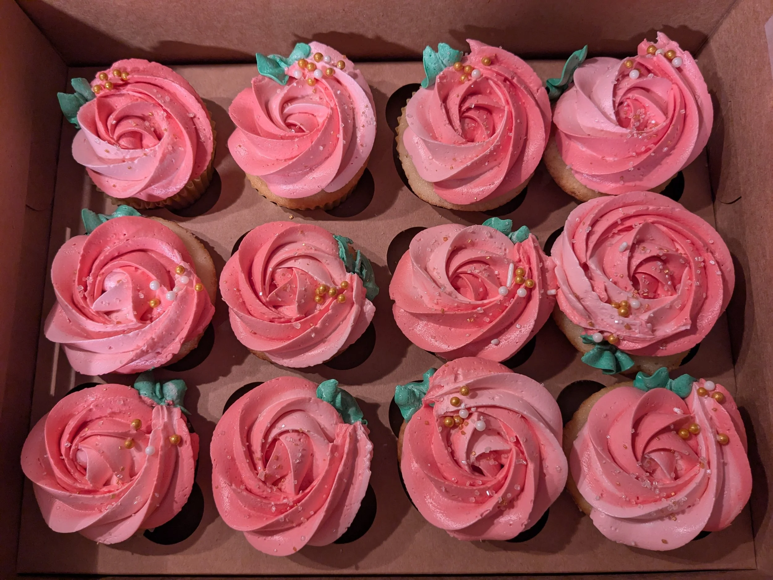 A box of twelve pink frosted cupcakes decorated with green leaves and small edible pearls.
