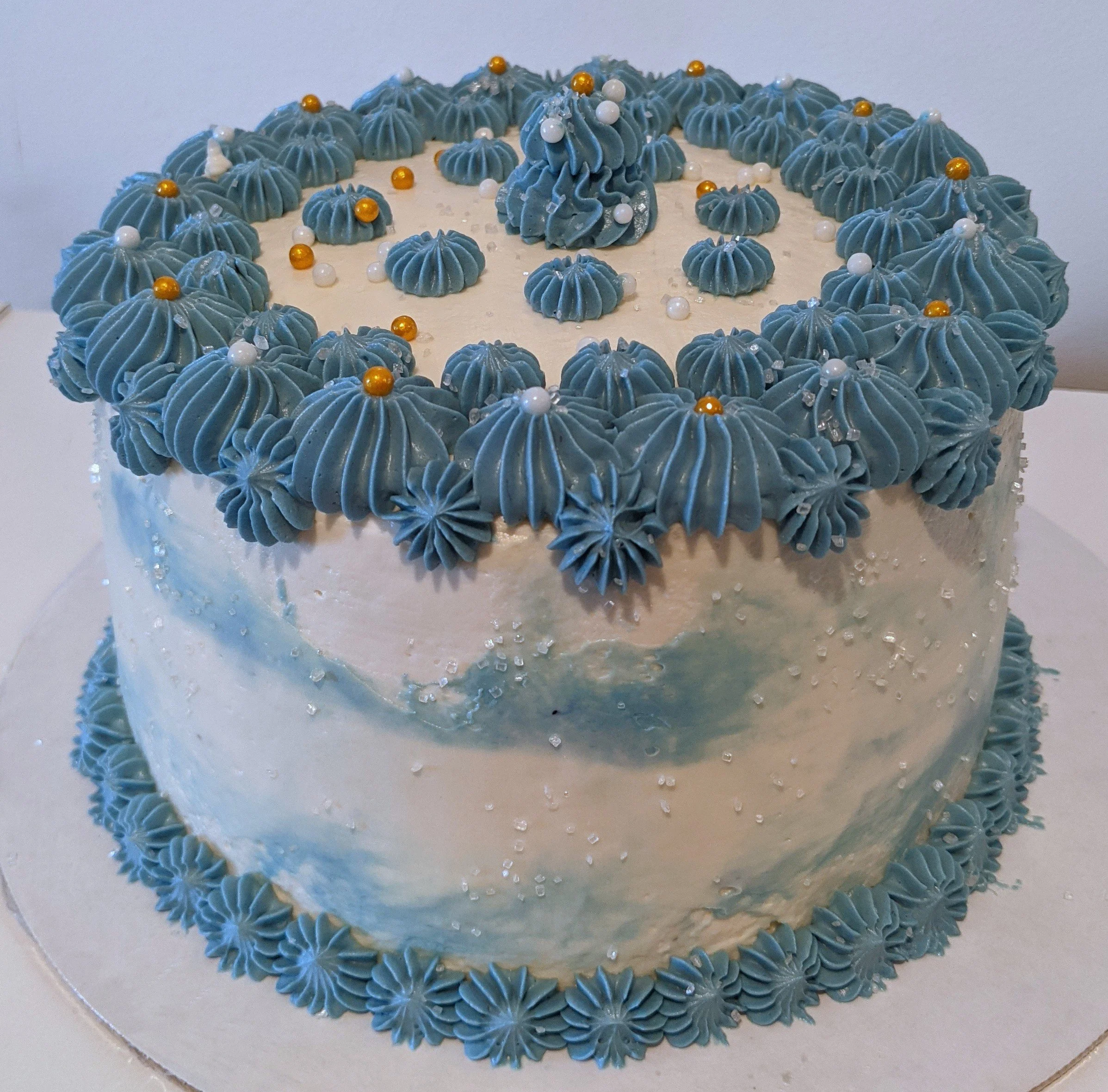 A round cake decorated with white and blue frosting, topped with blue frosting rosettes and pearl-like decorations, with a border of blue frosting rosettes at the base.