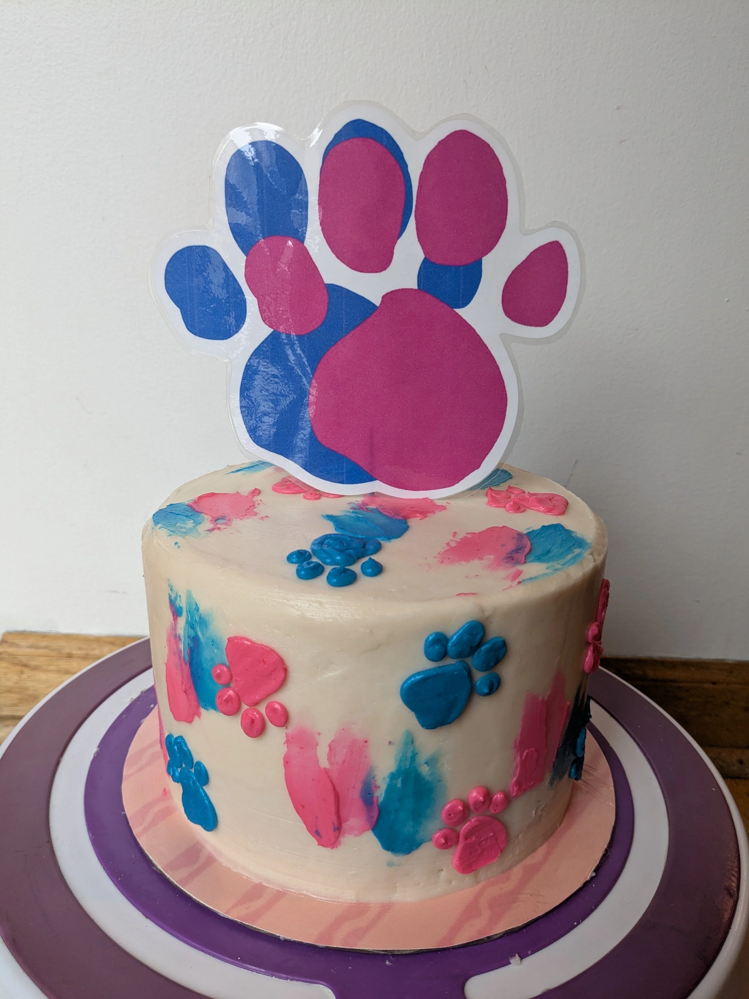 A white cake decorated with pink and blue paw prints and colorful brush strokes, topped with a large pink and blue paw print cutout on a stick.