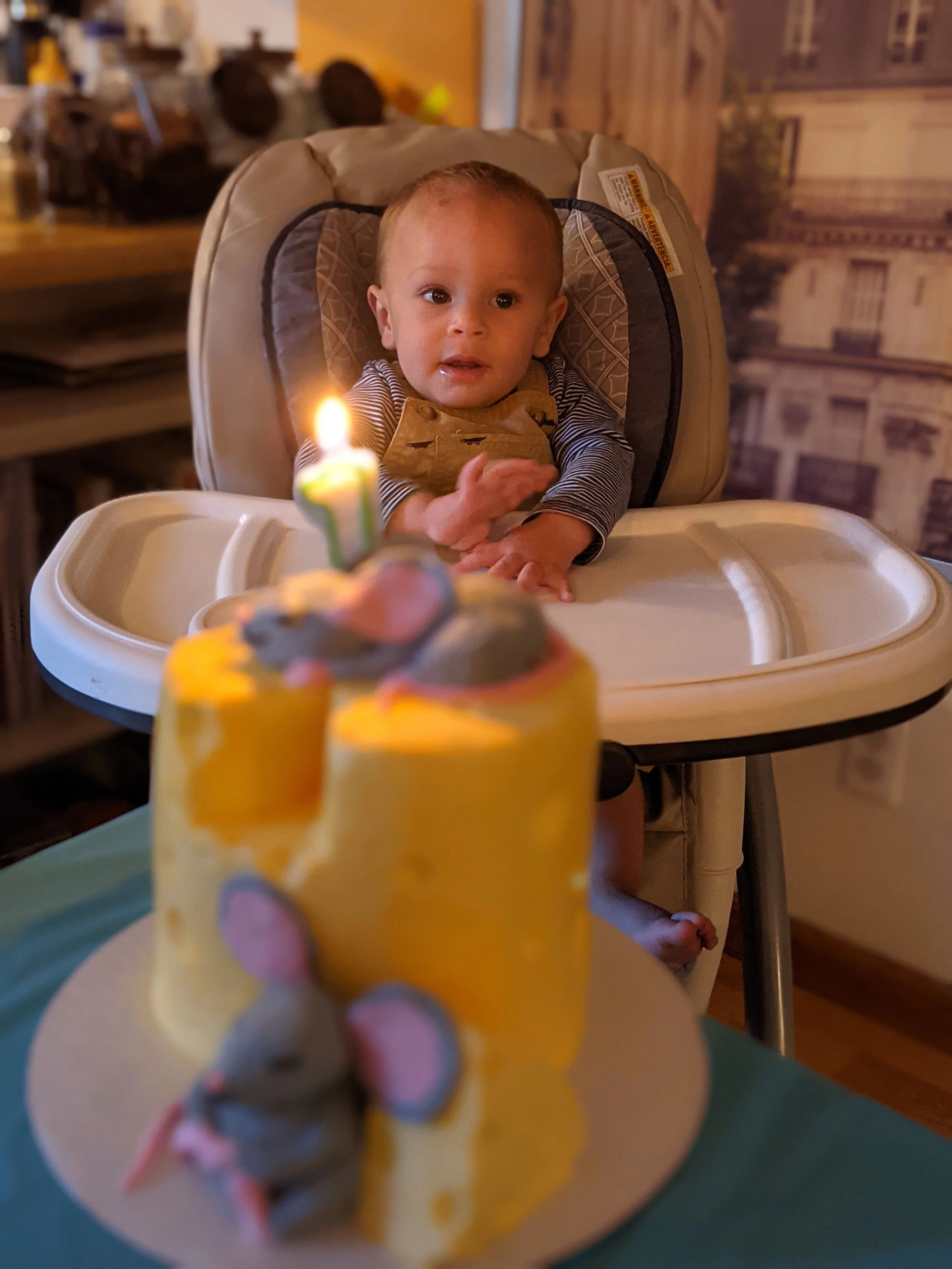 A young child sitting in a high chair looking at a small birthday cake with a lit candle featuring a mouse decoration and a mouse figurine cake topper.