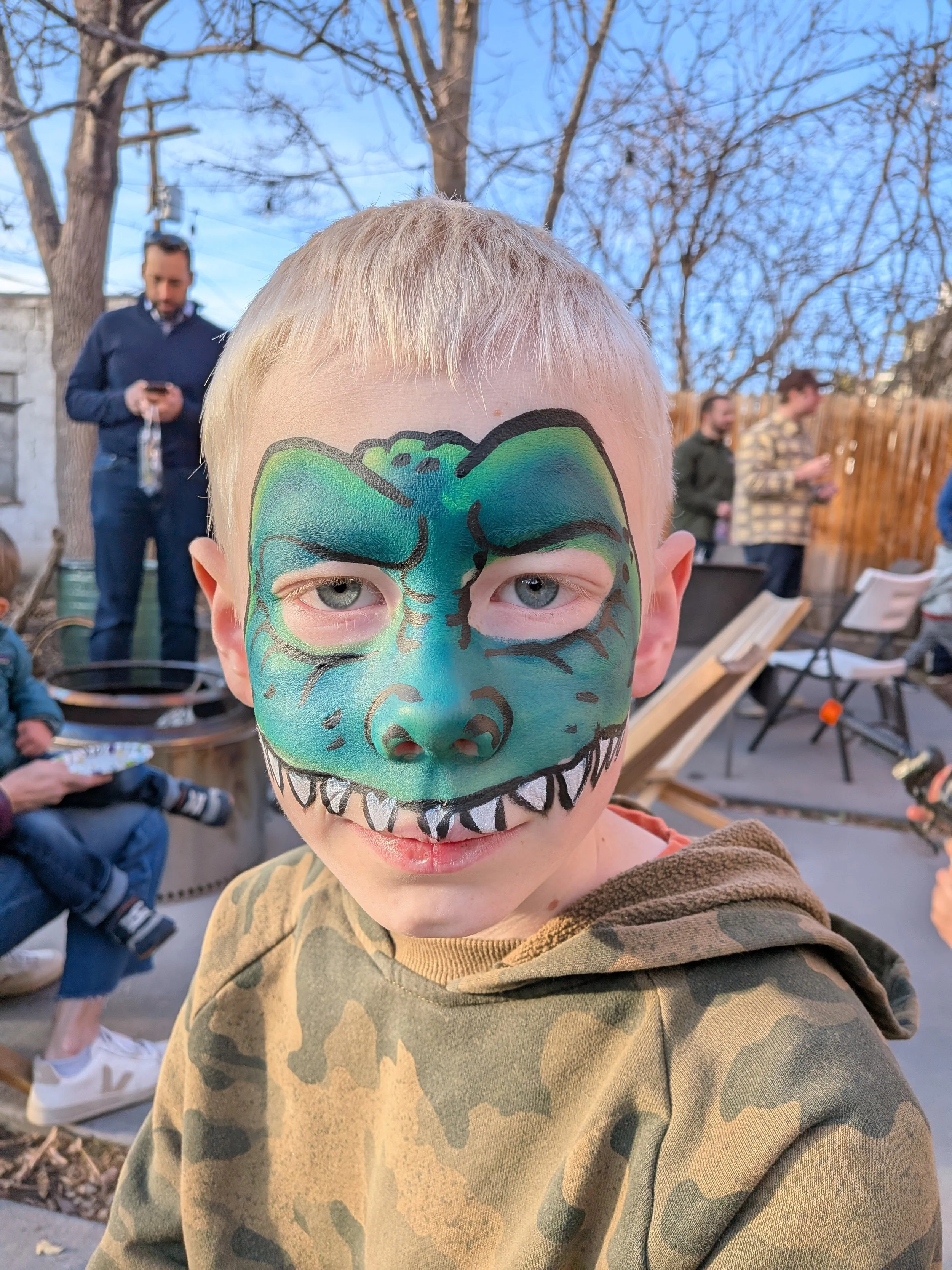 A young boy with face paint designed to look like a green dinosaur with sharp teeth, wearing a camouflage hoodie, outdoors during daytime with other people in the background.